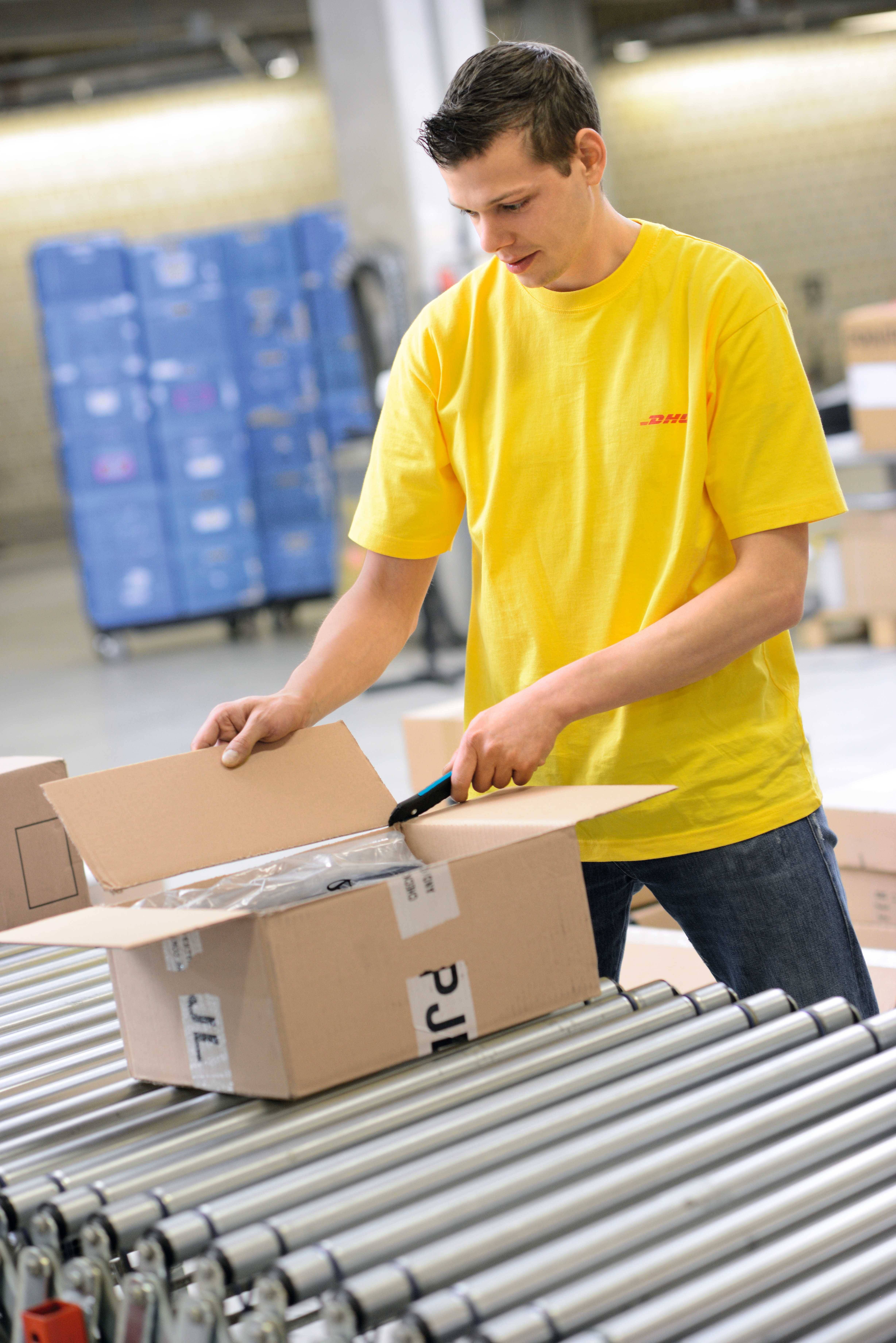 Un homme portant un t-shirt jaune ouvre un carton sur un tapis roulant dans un entrepôt. Des boîtes empilées sont visibles en arrière-plan.