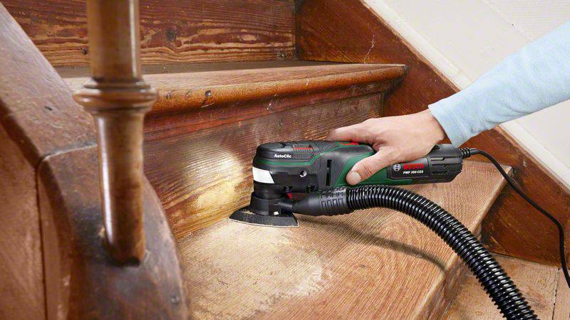 Une personne ponce un escalier en bois à l'aide d'un outil multifonction. L'outil est connecté à un aspirateur pour éliminer la poussière.