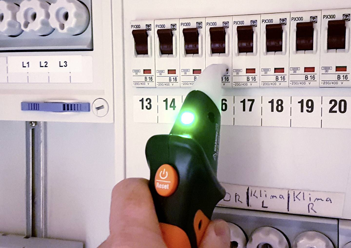Une main tient un testeur de tension avec une lumière verte allumée près d'un tableau électrique pour vérifier l'absence de tension.