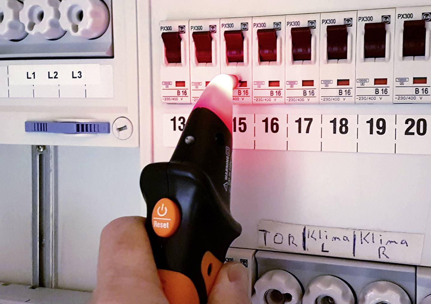 Une personne teste les fusibles dans un tableau électrique avec un testeur de tension qui s'allume en rouge au niveau du 16ème fusible.