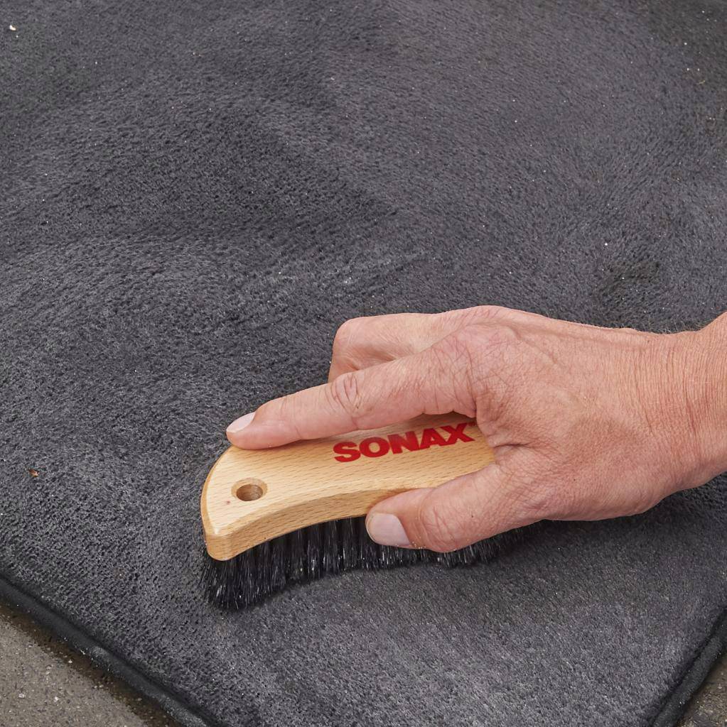 Une poignée en bois portant l'inscription 'SONAX' est tenue par une main. La brosse est utilisée pour nettoyer une surface grise.