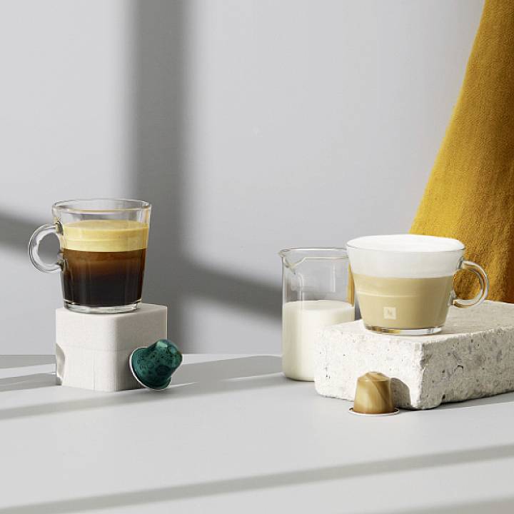 Deux tasses de café sur des pierres, accompagnées de deux capsules de café, l'une verte et l'autre dorée, dans un environnement minimaliste.