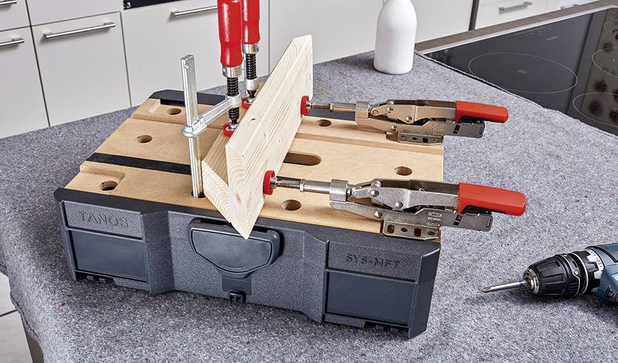Un établi avec un plateau en bois, maintenu par des serre-joints. Une perceuse est posée à côté. La scène montre une opération d'atelier.