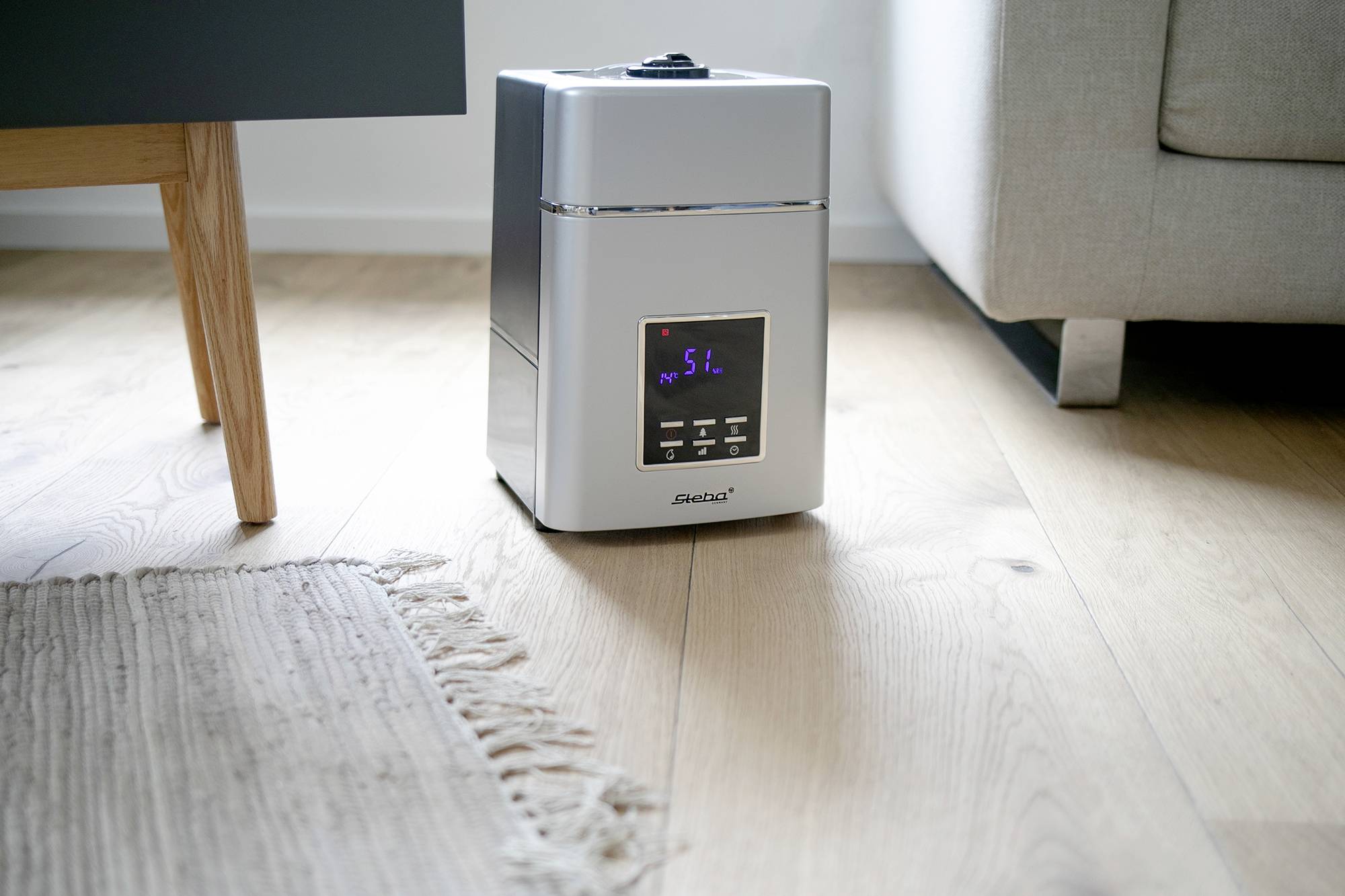 Un humidificateur est posé sur un plancher en bois à côté d'un tapis gris et d'un canapé. L'écran affiche « StL ».