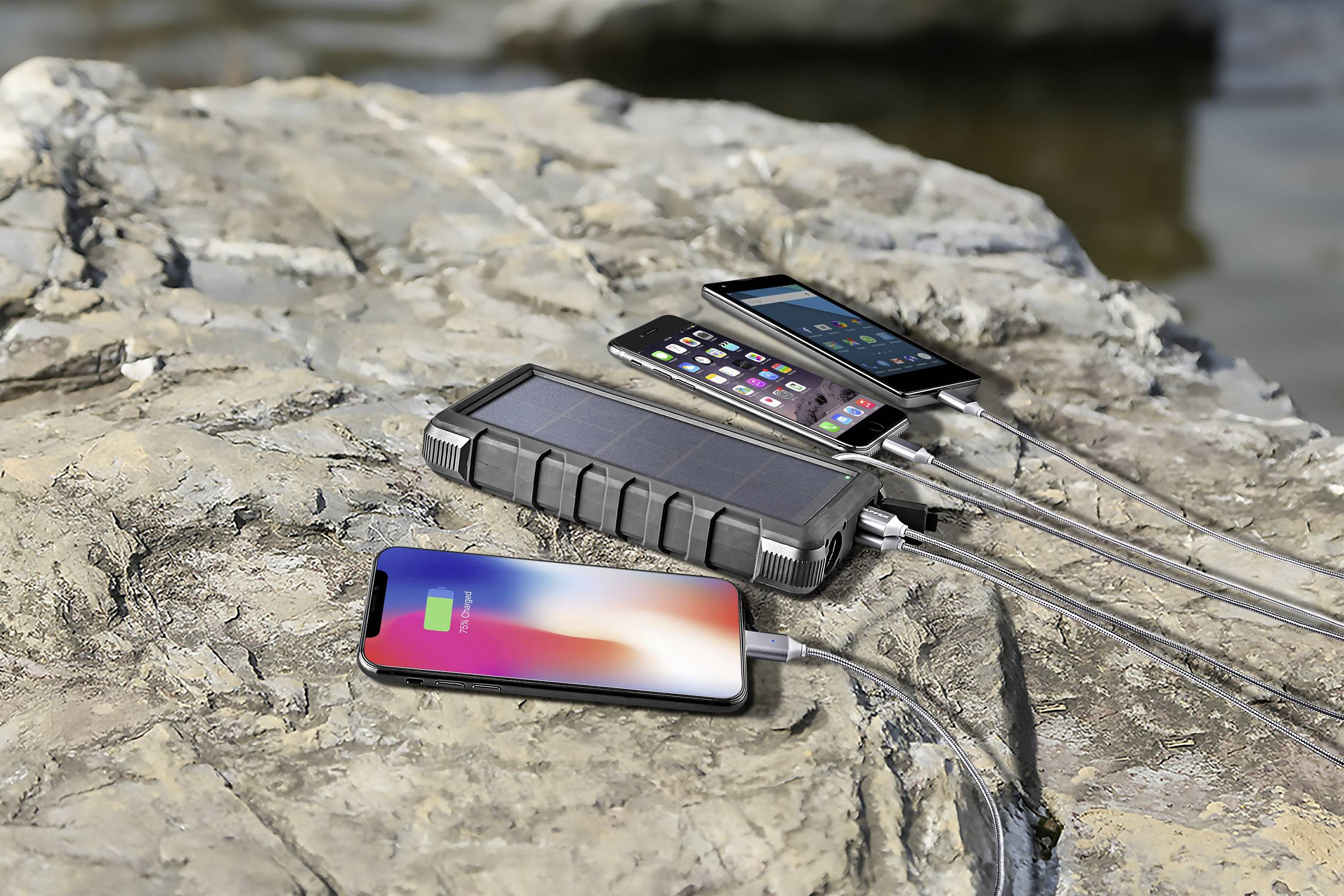 Une batterie solaire charge deux smartphones posés sur un grand rocher, avec une rivière visible en arrière-plan.