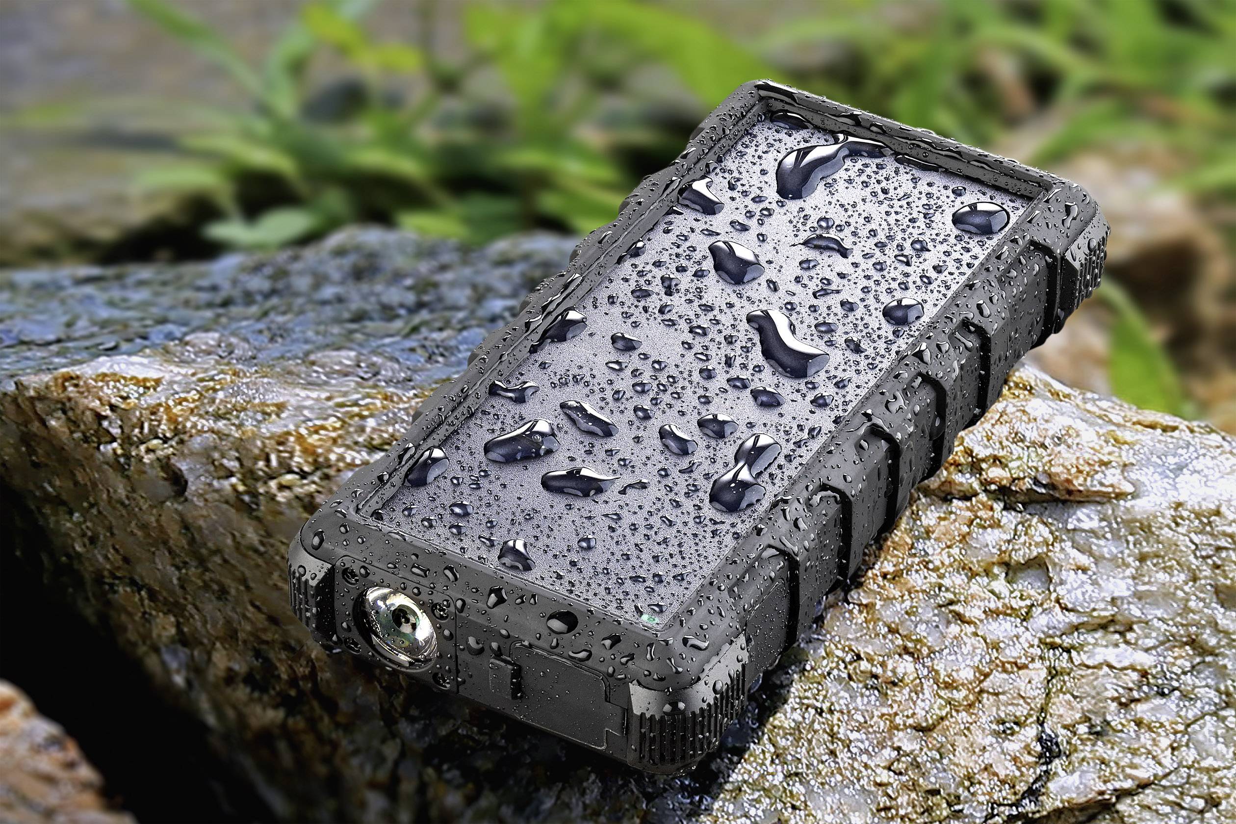 Une batterie externe robuste et étanche repose sur une pierre à l'extérieur, entourée de plantes. Elle est recouverte de gouttelettes d'eau.