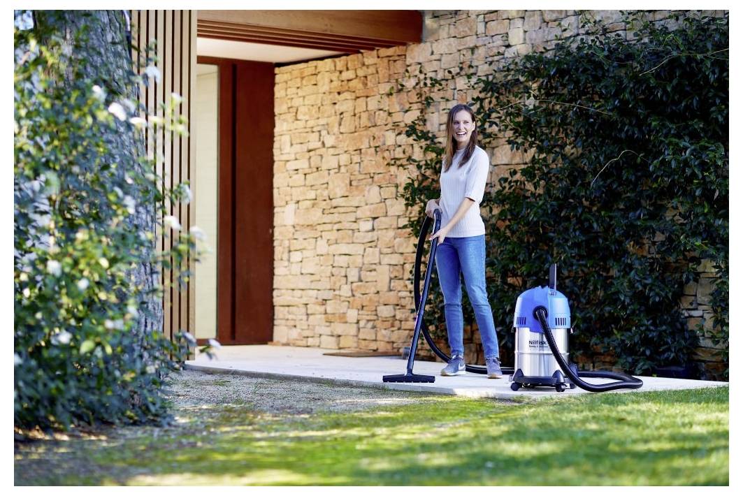 Une personne utilisant un aspirateur sur une terrasse près d'un mur en pierre et de la verdure, suggérant un nettoyage extérieur dans un environnement résidentiel.