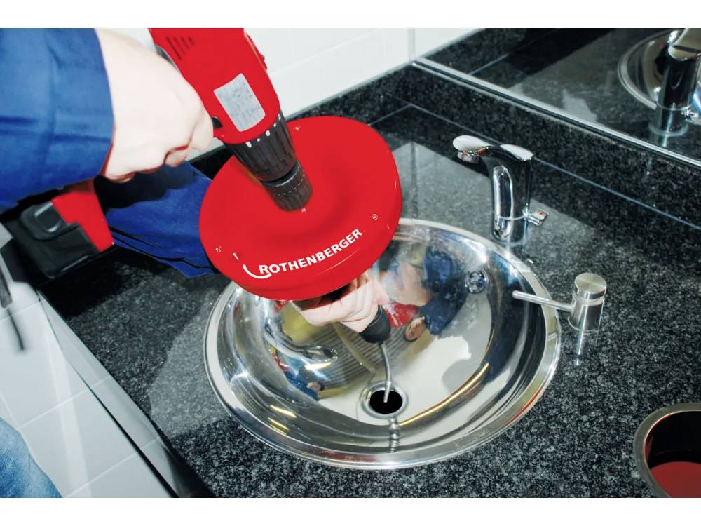 Une personne utilise un outil rouge Rothenberger pour déboucher un lavabo de salle de bain. L'outil est positionné au-dessus de la canalisation sur un plan de travail en granit.