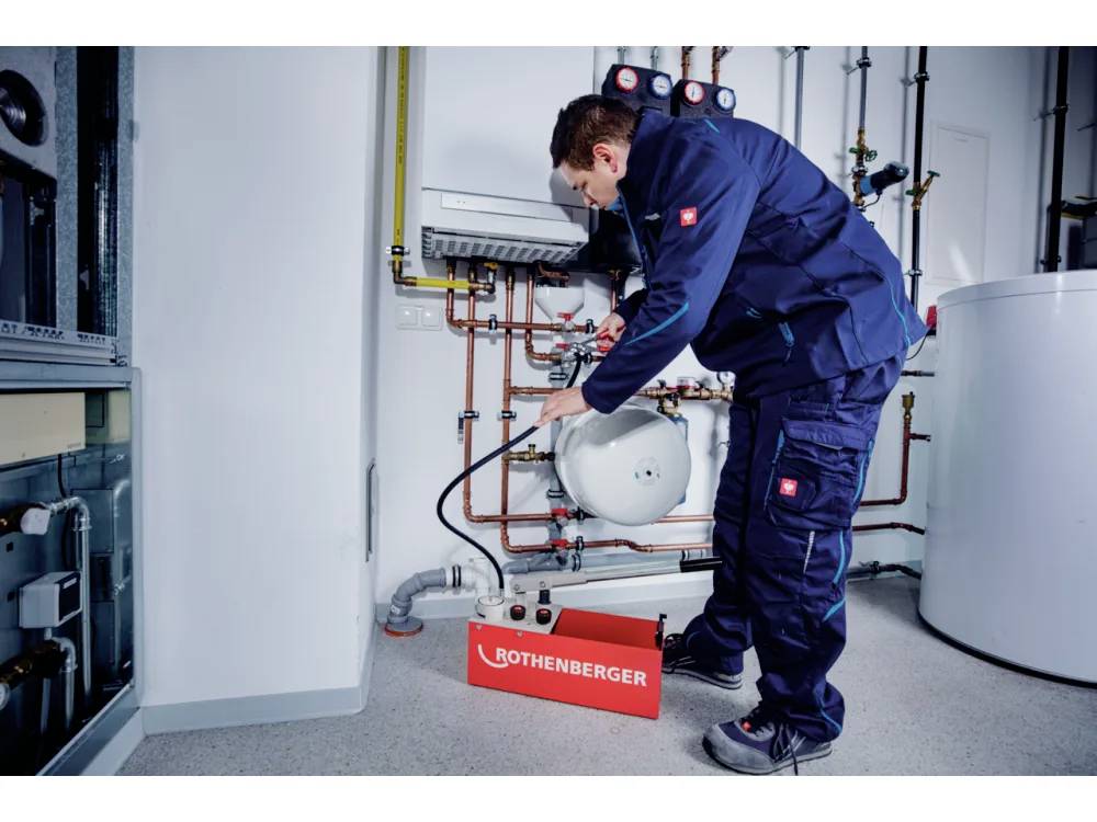 Un technicien en uniforme bleu utilise de l'équipement pour travailler sur des tuyaux dans une salle mécanique. Une boîte à outils Rothenberger rouge est posée au sol.