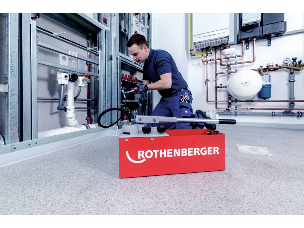Une personne travaille sur le système de tuyauterie d'un bâtiment, utilisant un outil 'Rothenberger' posé au sol. Des tuyaux industriels et de l'équipement l'entourent.