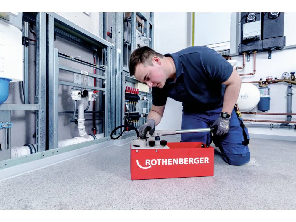Une personne utilise un outil d'un coffret rouge 'ROTHENBERGER' dans un local technique avec divers tuyaux et équipements sur les murs.