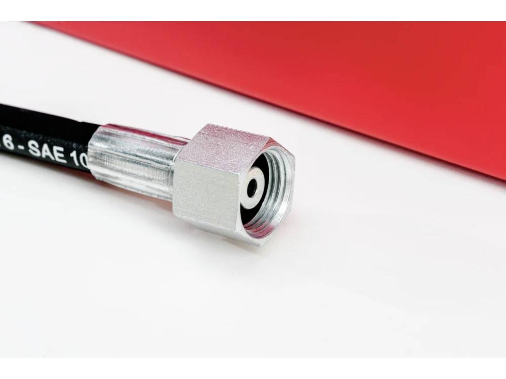 Un raccord de tuyau hydraulique métallique fixé à un tuyau noir repose sur une surface blanche et rouge, indiquant la connectivité d'un équipement industriel.