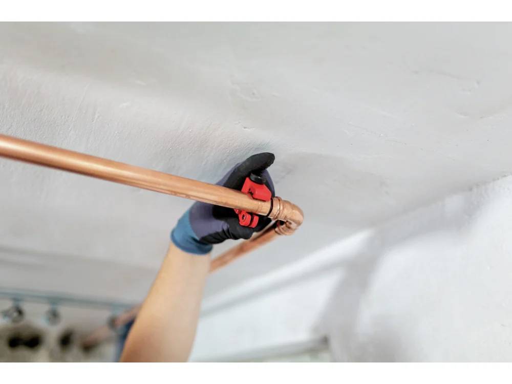 Une personne portant des gants serre un tuyau en cuivre sous un plafond blanc à l'aide d'un outil rouge, suggérant une tâche de plomberie ou de maintenance.