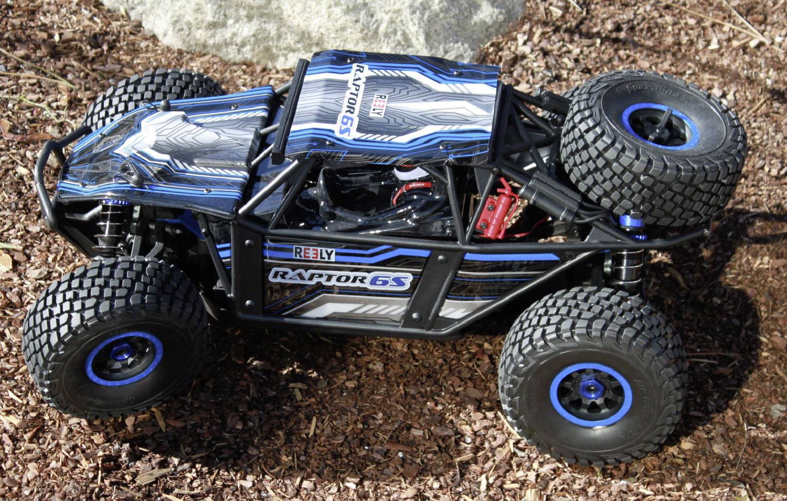 Un buggy RC avec l'inscription 'Raptor 6S' et 'Arrma' en bleu et noir, est posé sur terre. La voiture a de grandes roues au profil très marqué.