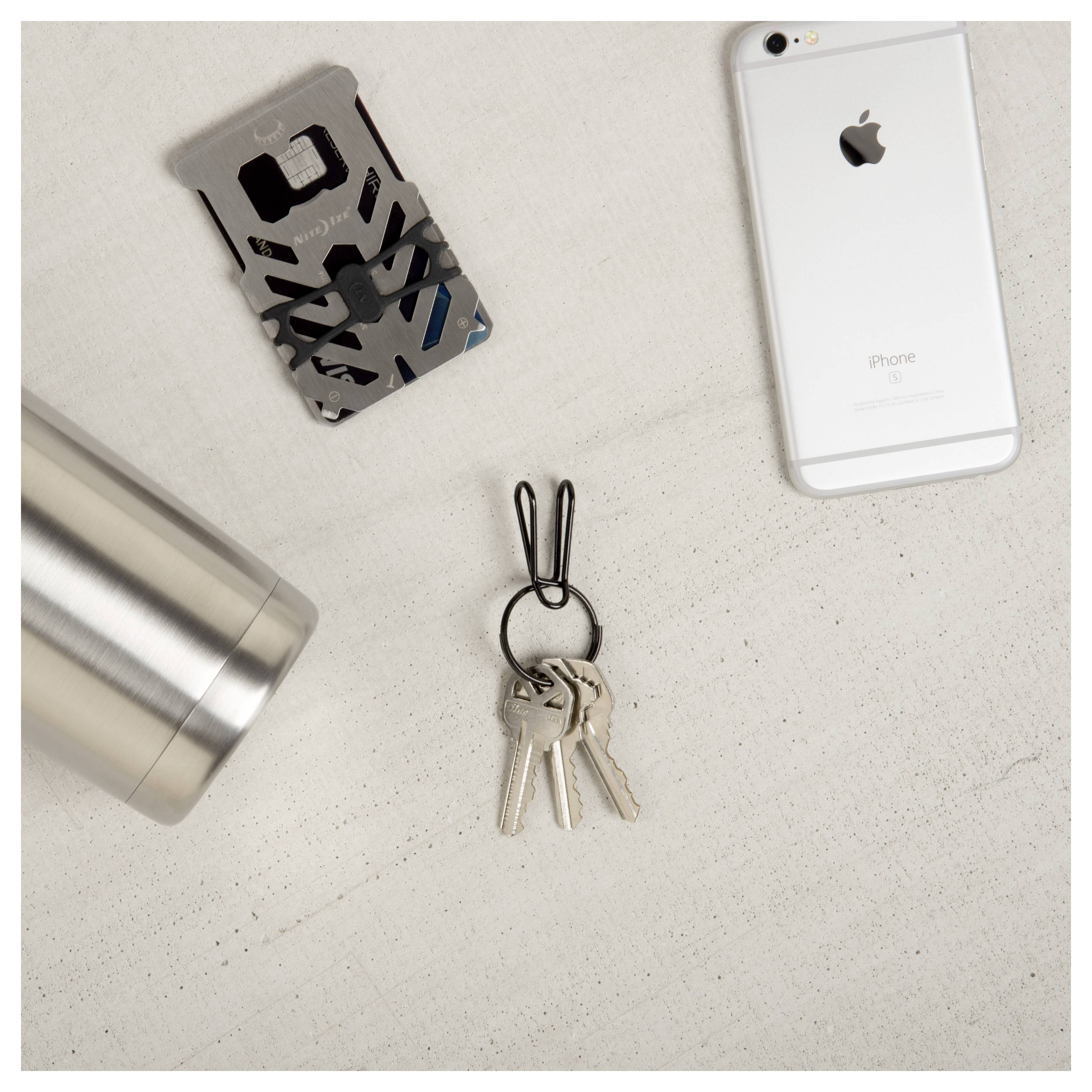 Un smartphone, un gobelet en métal, une carte multifonction et un porte-clés avec des clés sont disposés sur une surface claire.