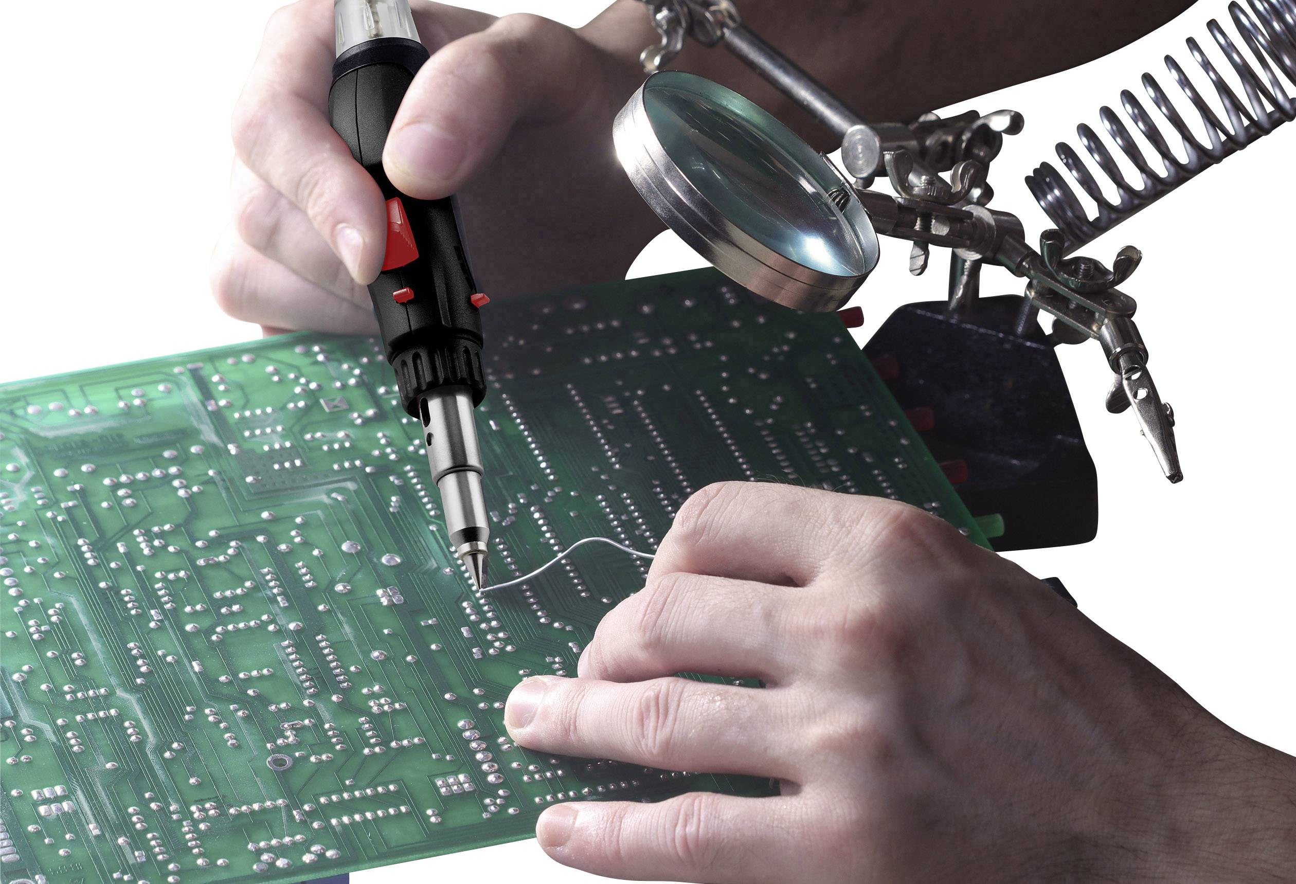 Gros plan de mains travaillant avec un fer à souder sur une carte de circuit imprimé verte, avec une loupe et une pince pour stabiliser le travail.