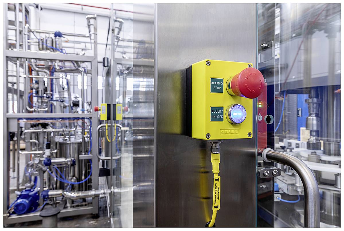 Un bouton d'arrêt d'urgence jaune sur une machine industrielle dans une halle d'usine, avec des tuyauteries complexes et des réservoirs en arrière-plan.