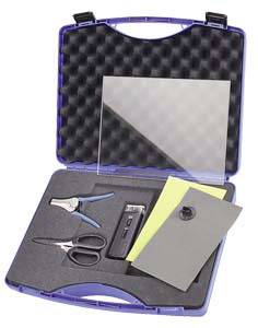 Une valise ouverte avec un intérieur en mousse, contenant une pince, des ciseaux, une feuille rectangulaire transparente, une feuille jaune et un objet gris.