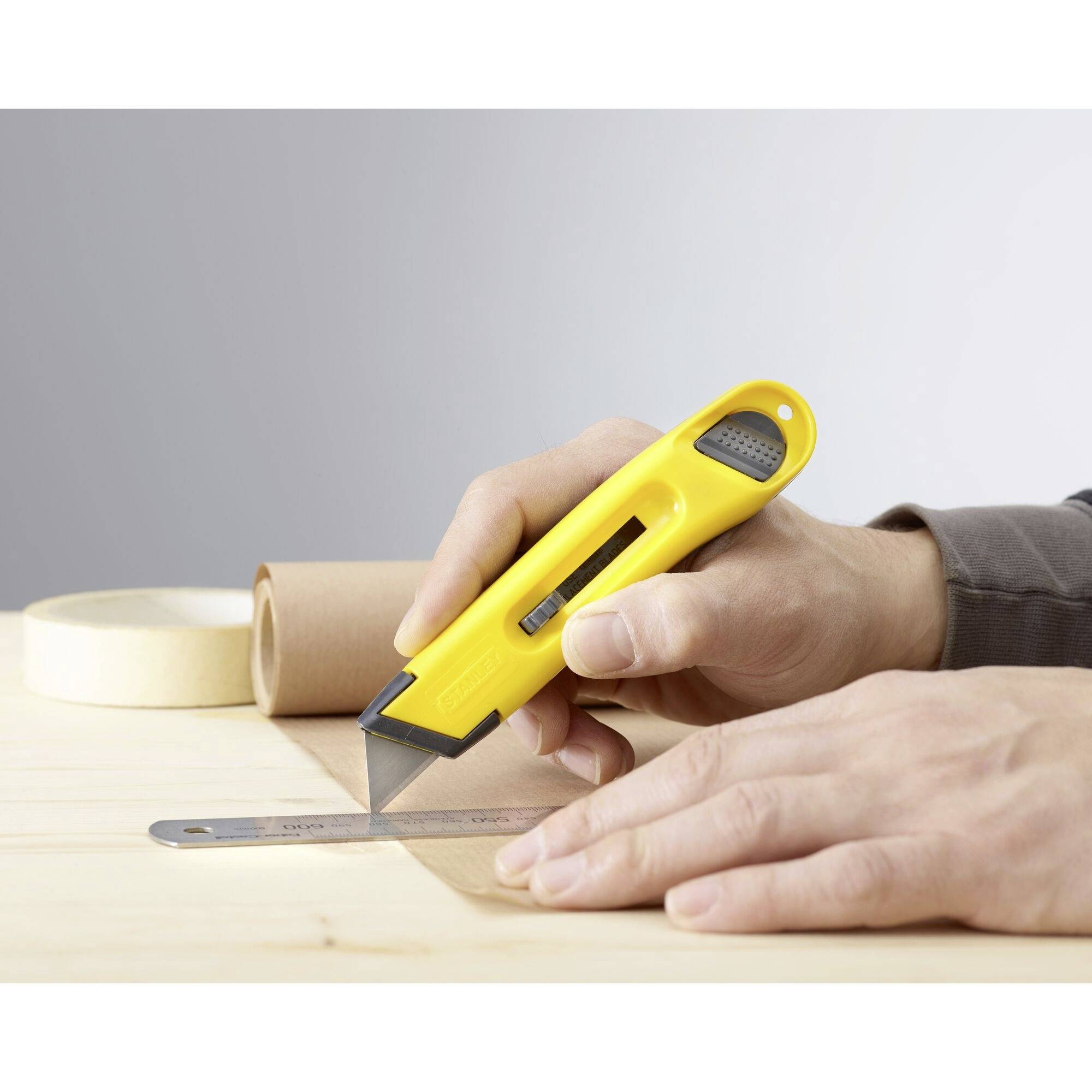 Une personne coupe avec un cutter jaune le long d'une règle métallique sur une table en bois. Des rouleaux de ruban adhésif sont visibles en arrière-plan.