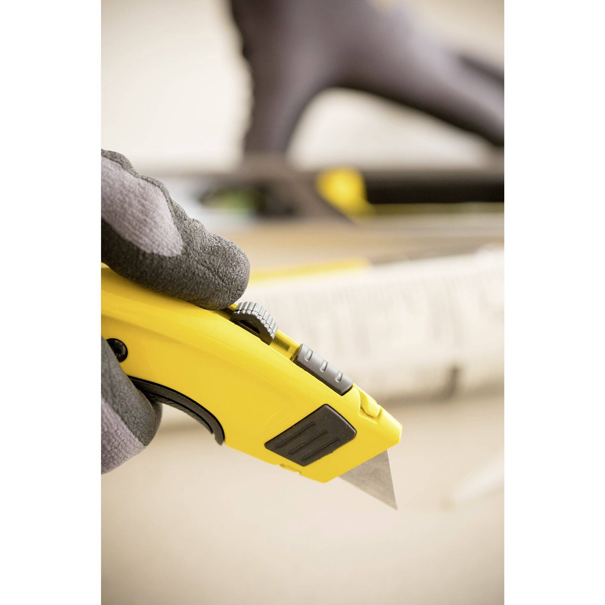 Une main tient un cutter jaune utilisé pour couper du carton. L'arrière-plan est flou, avec la mise au point sur le couteau.