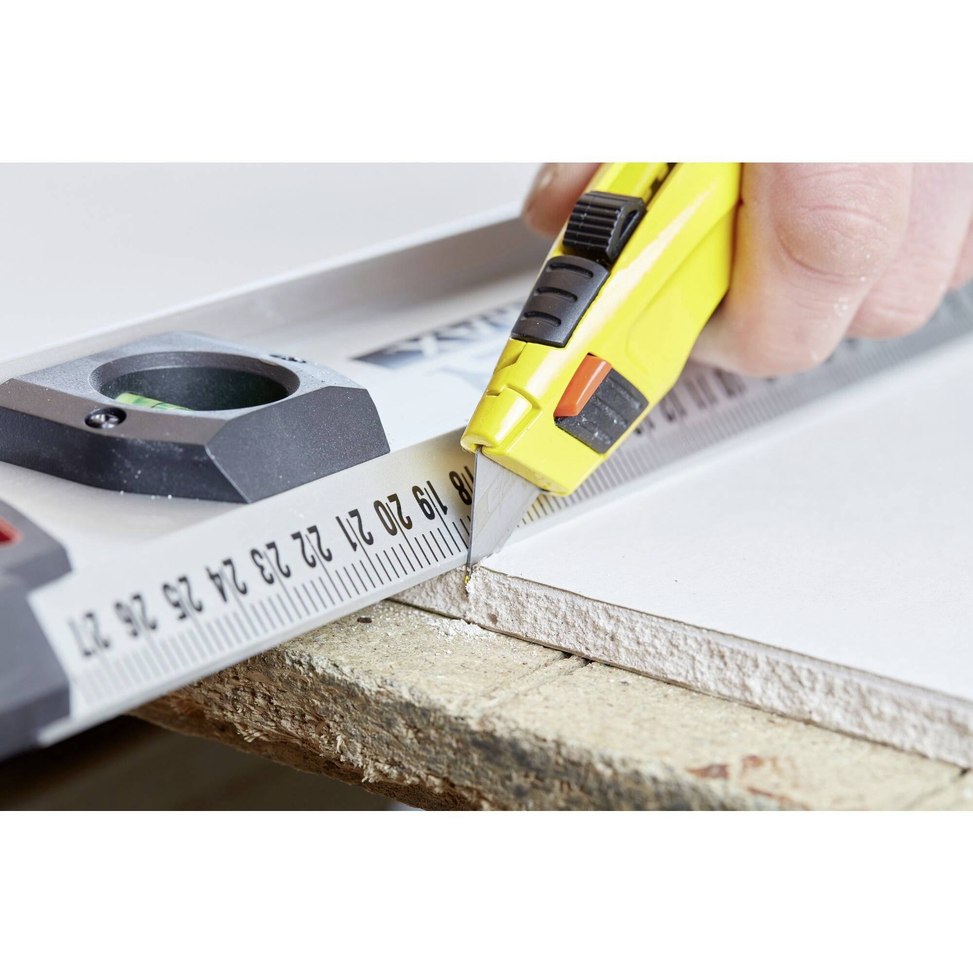 Une main utilise un cutter jaune et une règle sur une planche en bois pour une coupe précise.
