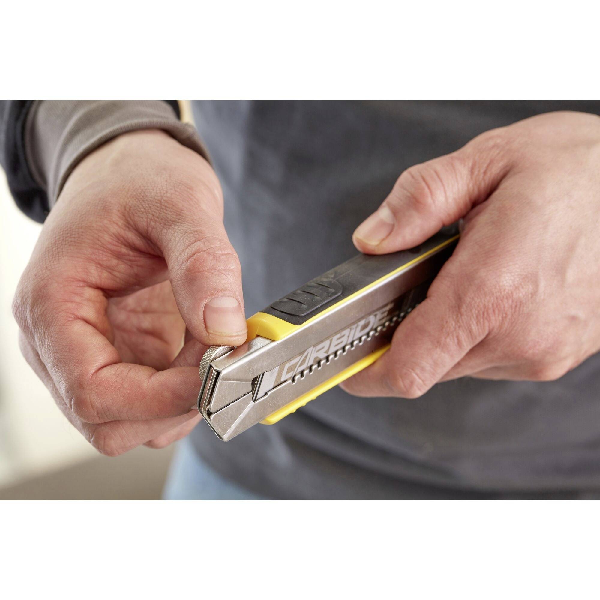 Une personne tient un couteau à tapis argent et noir dans ses mains et tourne la pointe pour le fixer ou le retirer.