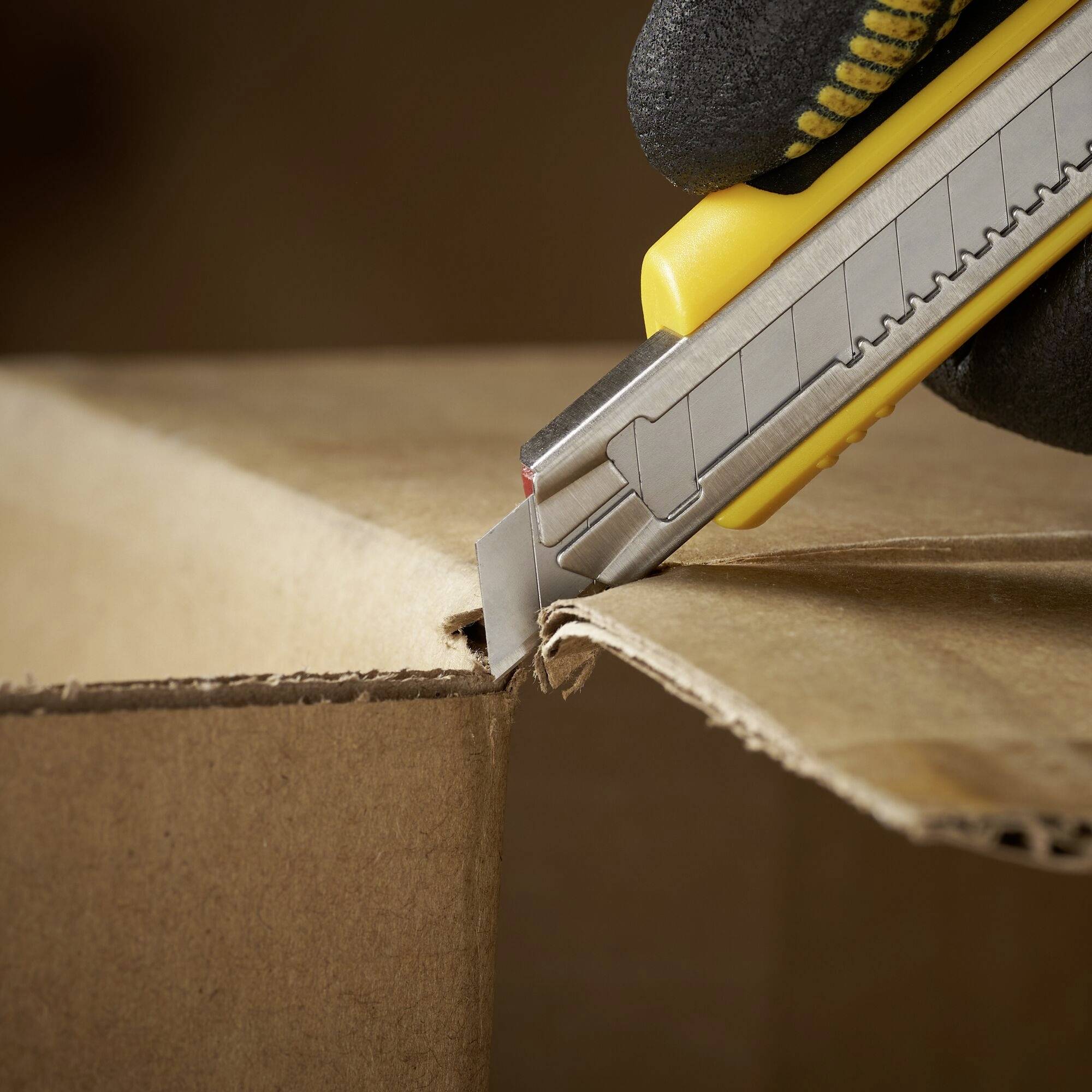 Une main tenant un cutter jaune coupe soigneusement un carton le long de son bord.