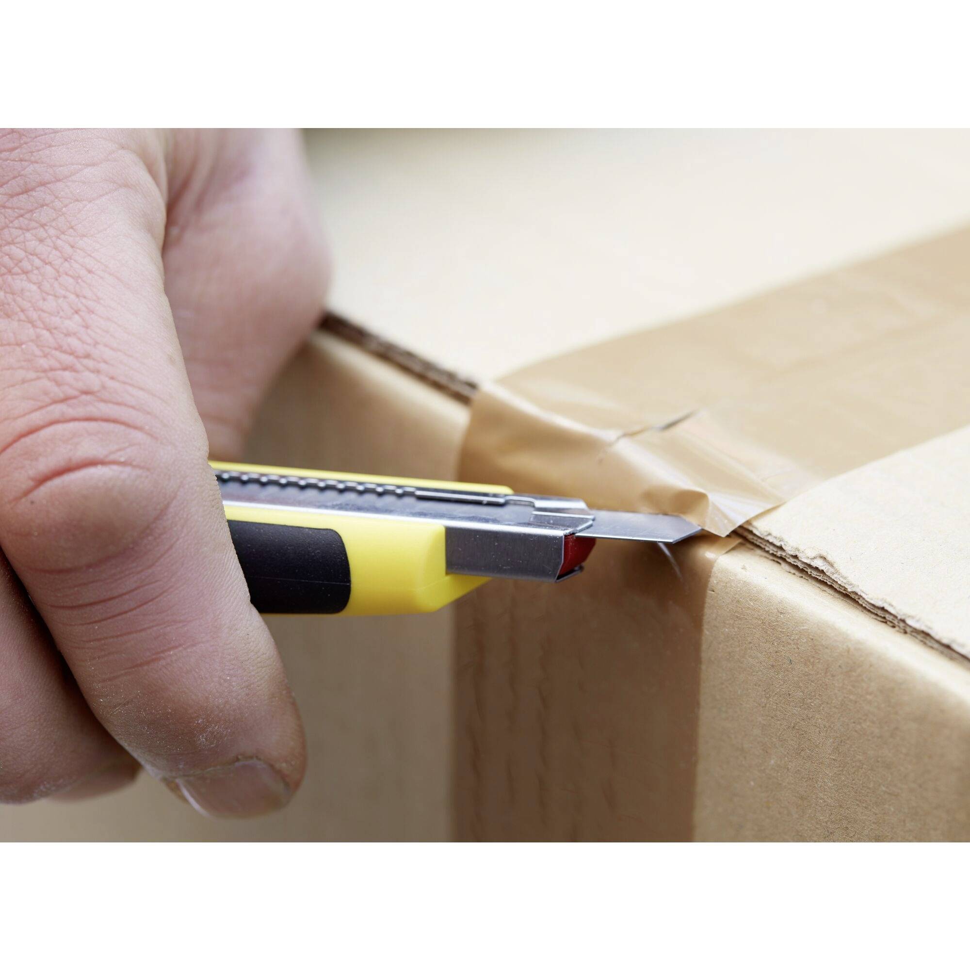 Une main coupe du ruban adhésif sur un carton à l'aide d'un cutter jaune.