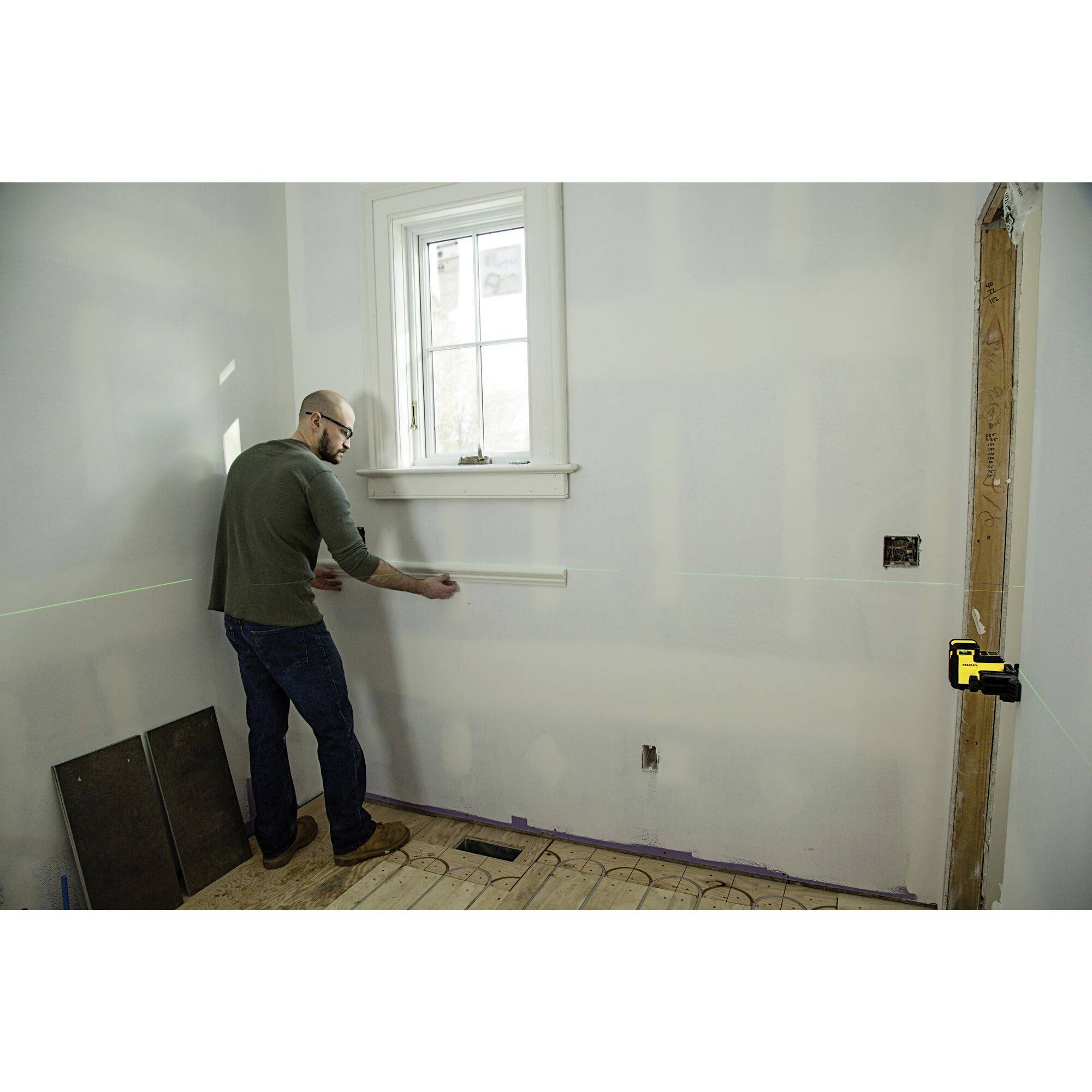 Un homme mesure un mur à l'aide d'un mètre ruban dans une pièce rénovée. Des lignes laser sont tracées sur le mur pour un alignement précis.