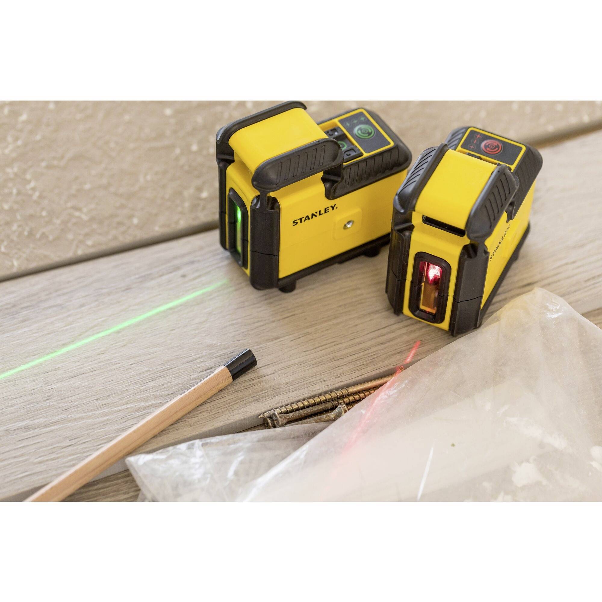 Télémètre laser et mesureur de vitesse sur un plancher en bois, ligne verte projetée. À côté, des vis, un crayon et un sac en plastique.
