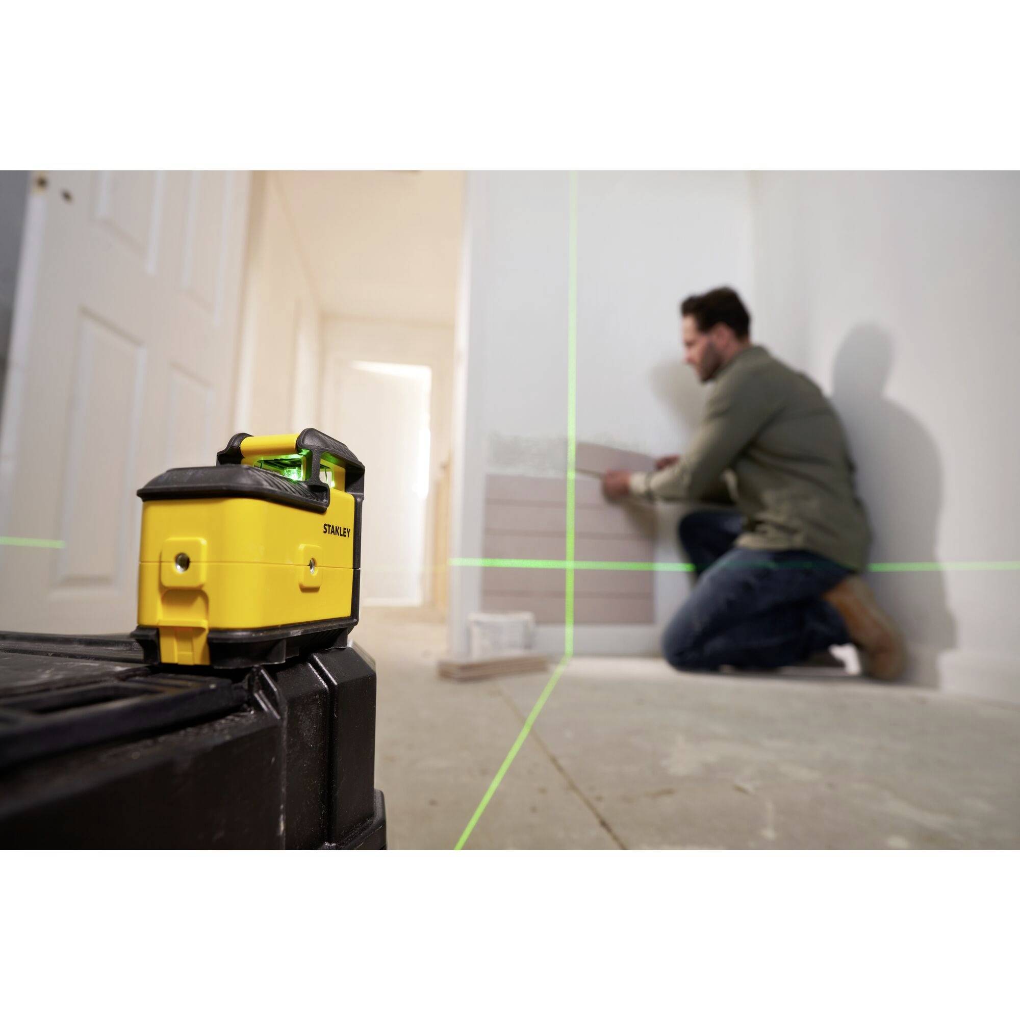 Un homme utilise un niveau laser pour poser des carreaux sur un mur. L'appareil projette des lignes vertes pour un alignement précis.