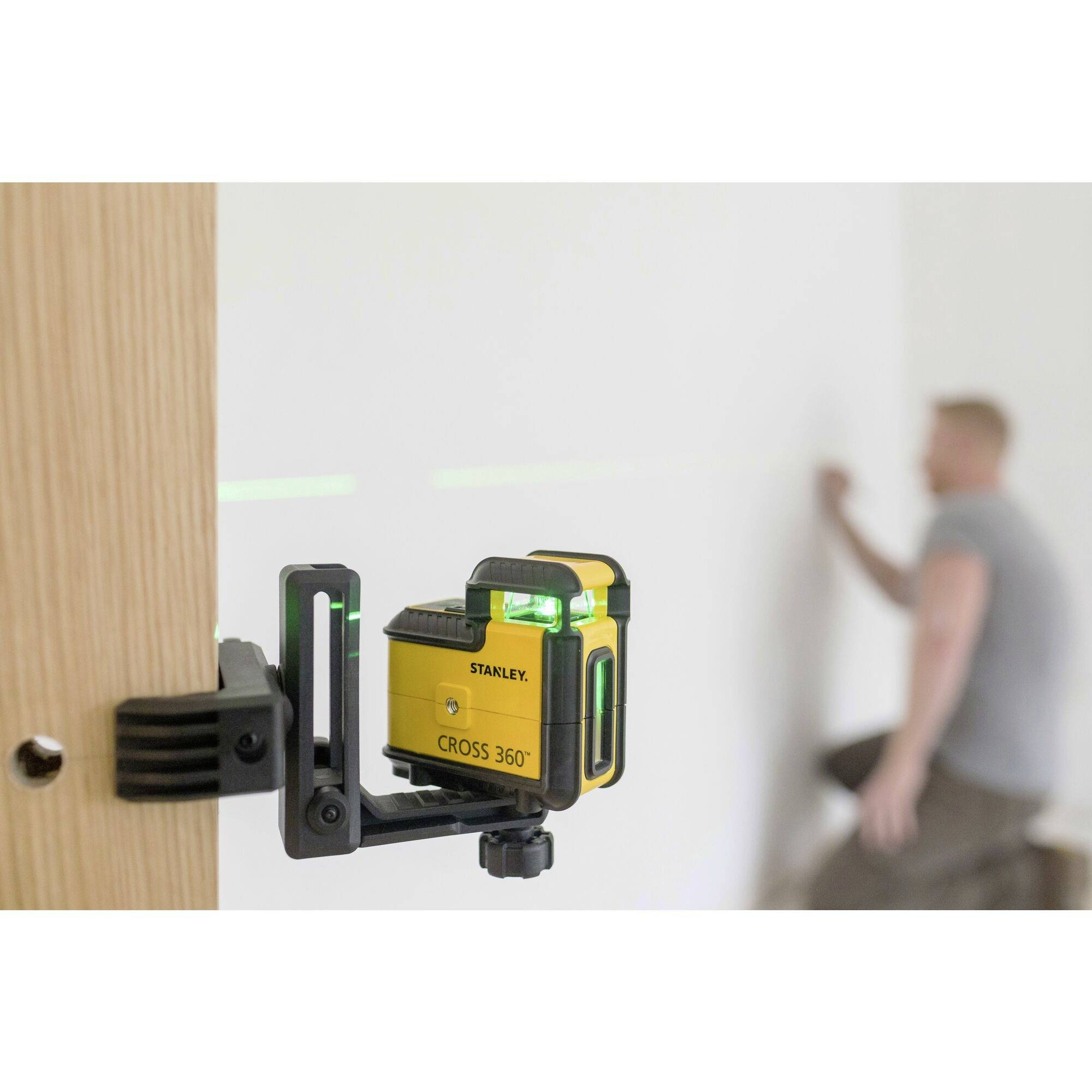 Un laser à lignes croisées est fixé à un morceau de bois et projette des lignes vertes sur un mur blanc. En arrière-plan, une personne floue travaille près du mur.