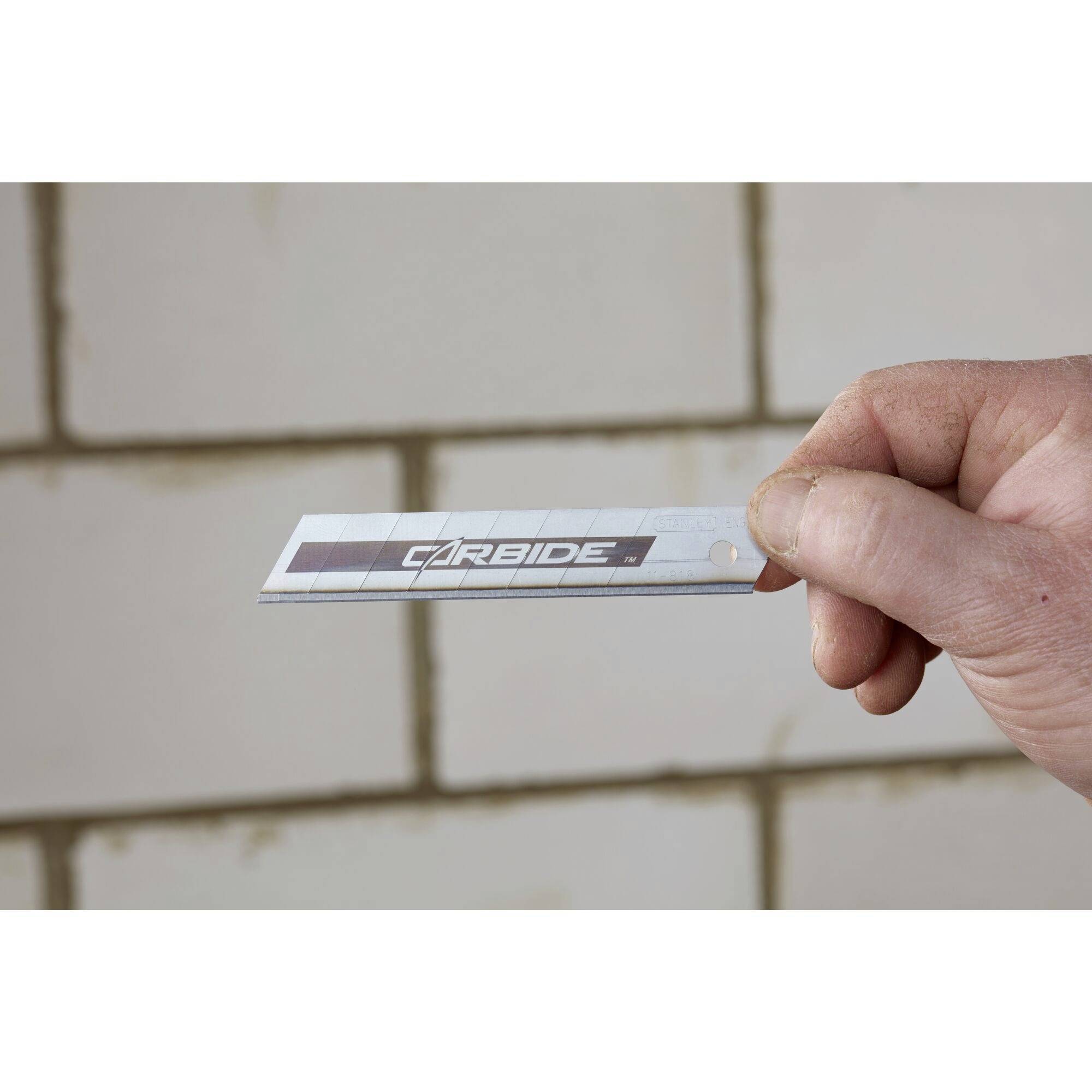 Une main tient une lame étroite portant l'inscription 'CARBIDE' devant un mur de carreaux clairs.