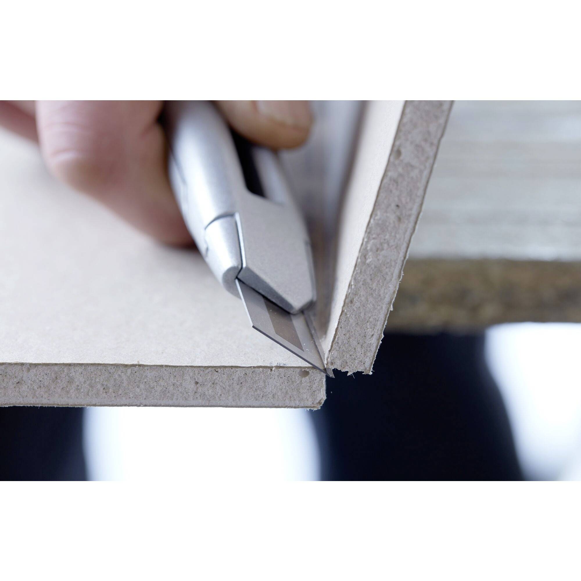 Une main coupe une ligne droite dans une plaque de carton à l'aide d'un cutter, posée sur une table.