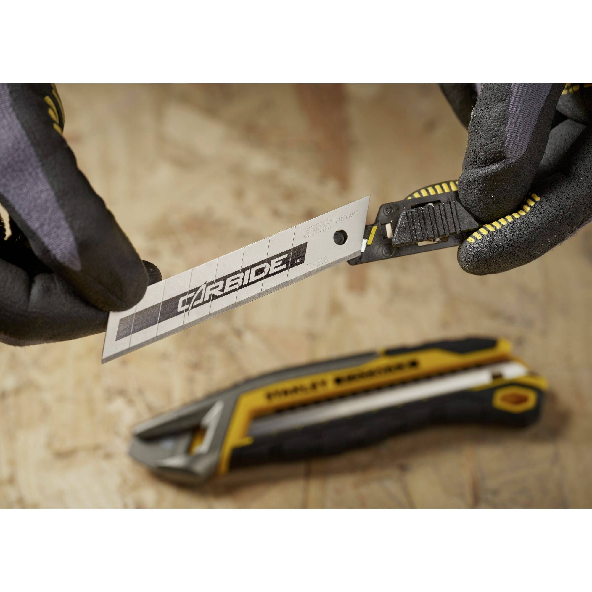 Deux mains gantées tiennent une lame portant l'inscription 'Carbide', avec un cutter jaune en arrière-plan sur un support en bois.