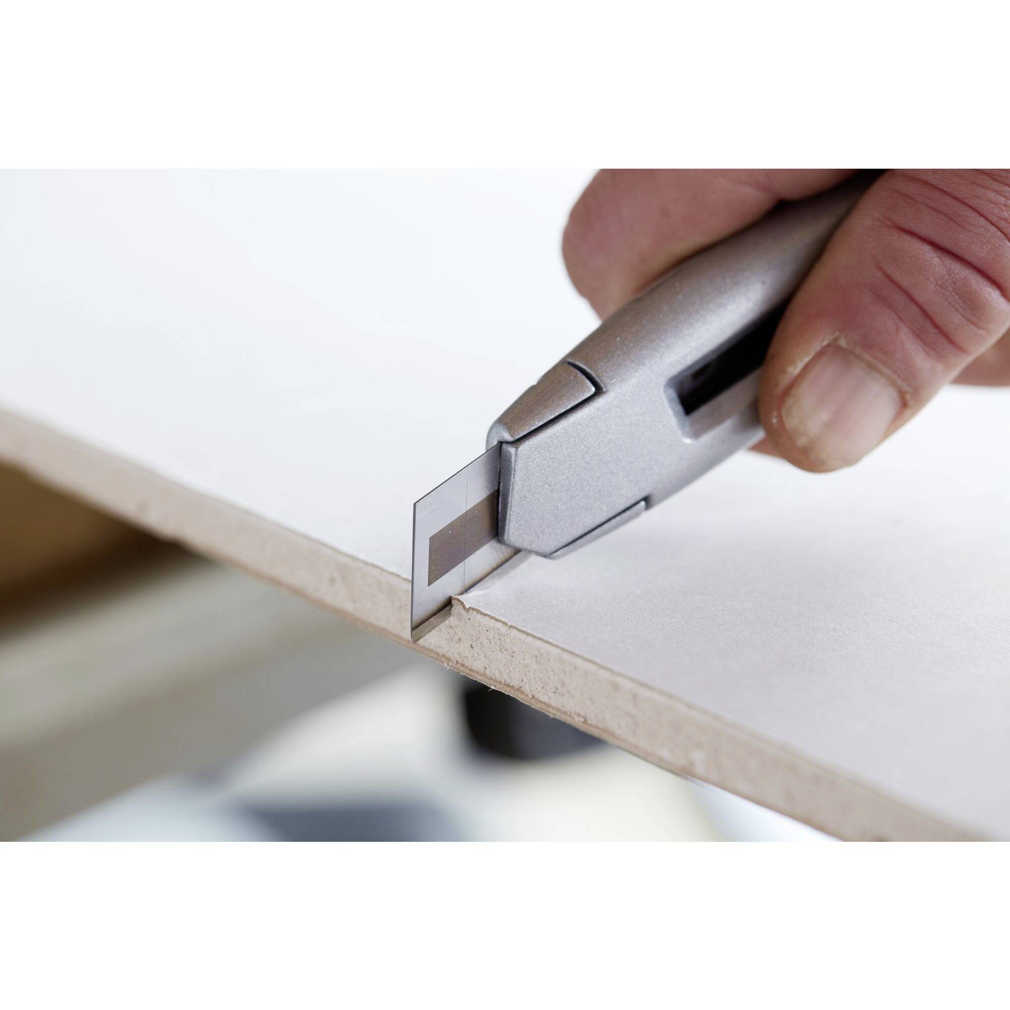Une main trace des lignes de précision sur un carton blanc à l'aide d'un couteau universel argenté, le long du bord d'une table.