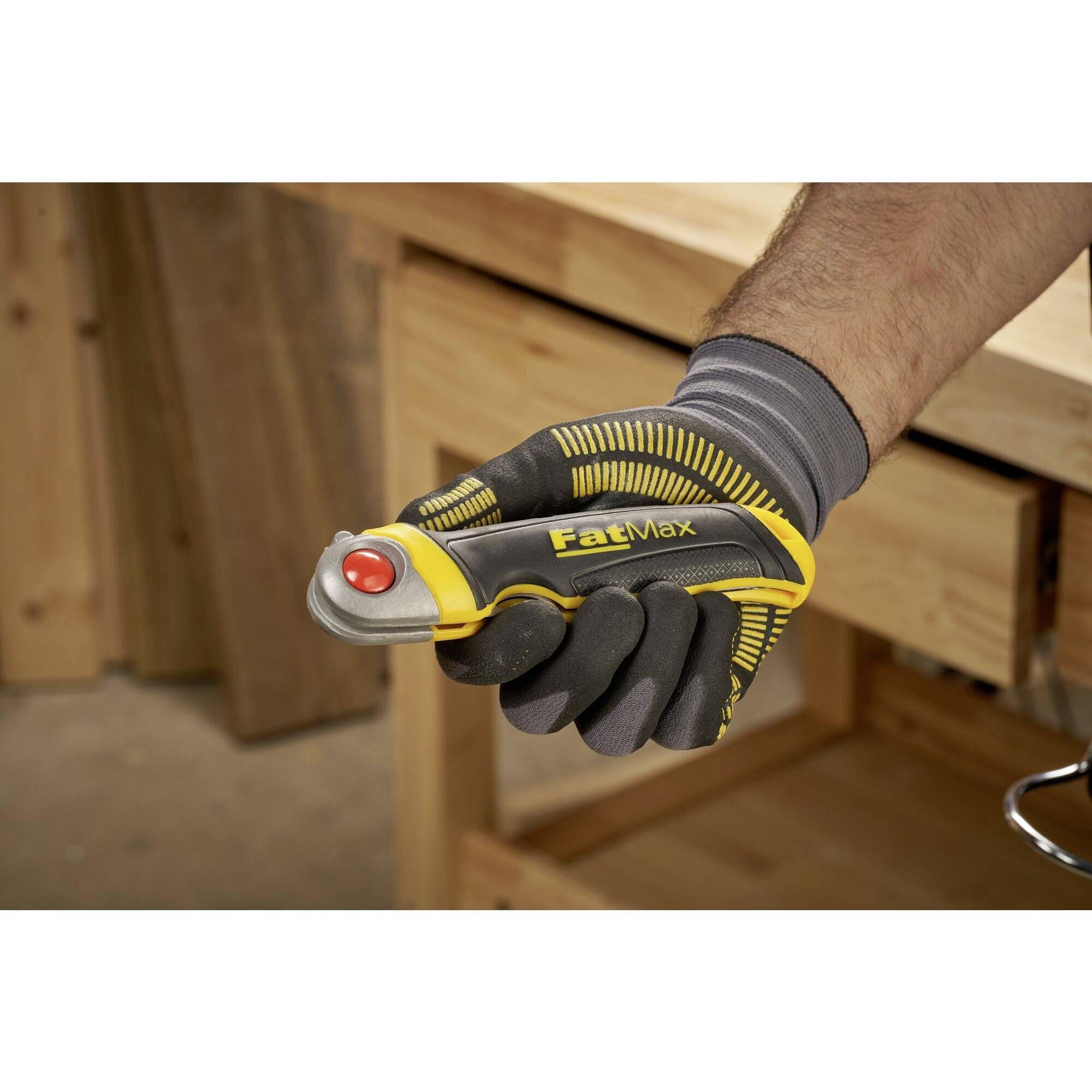 Une main portant un gant jaune et noir tient un couteau de sécurité FatMax jaune avec un bouton rouge devant un établi.