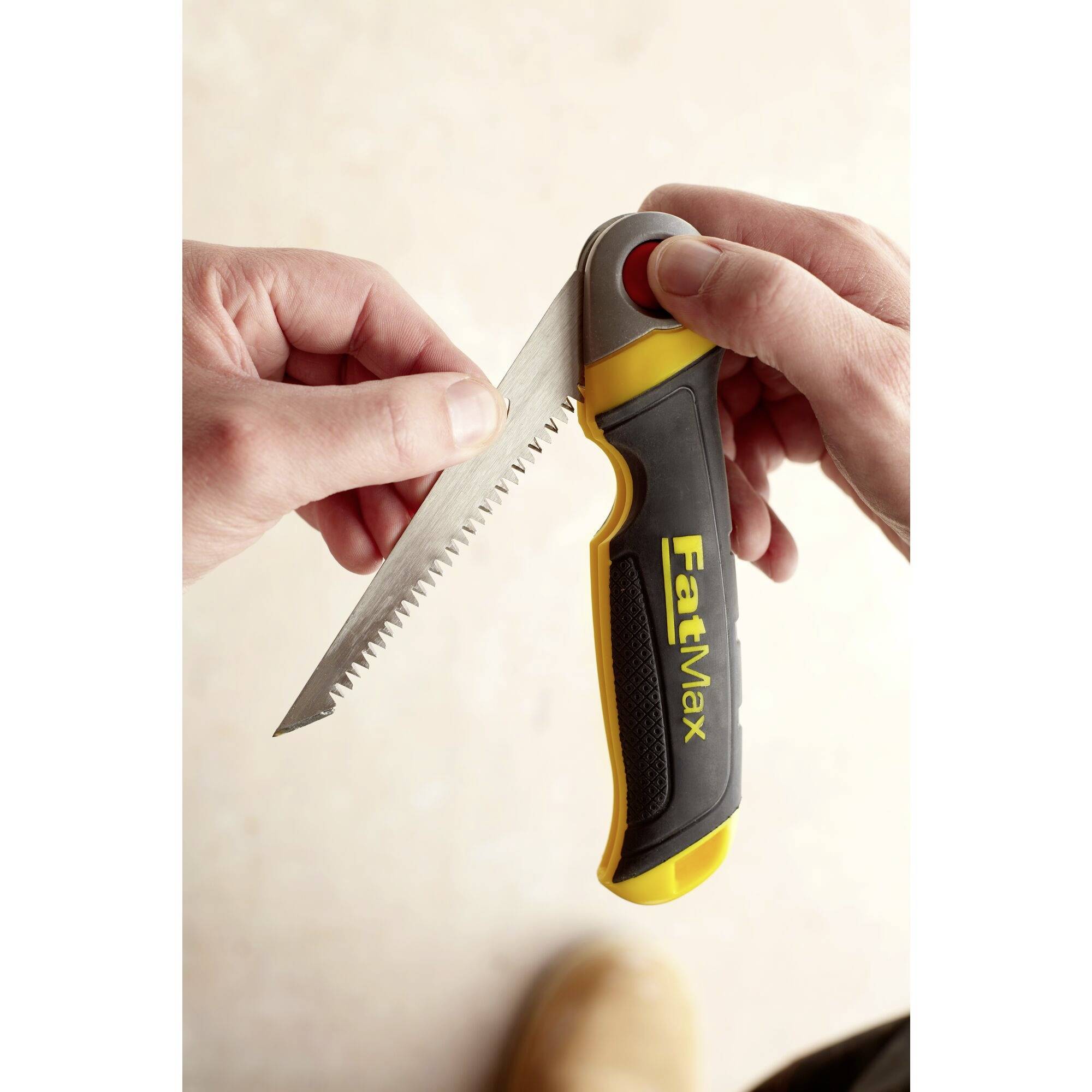 Deux mains tiennent une scie à main pliante dentelée avec une poignée jaune. L'inscription 'FatMax' est visible sur la poignée.