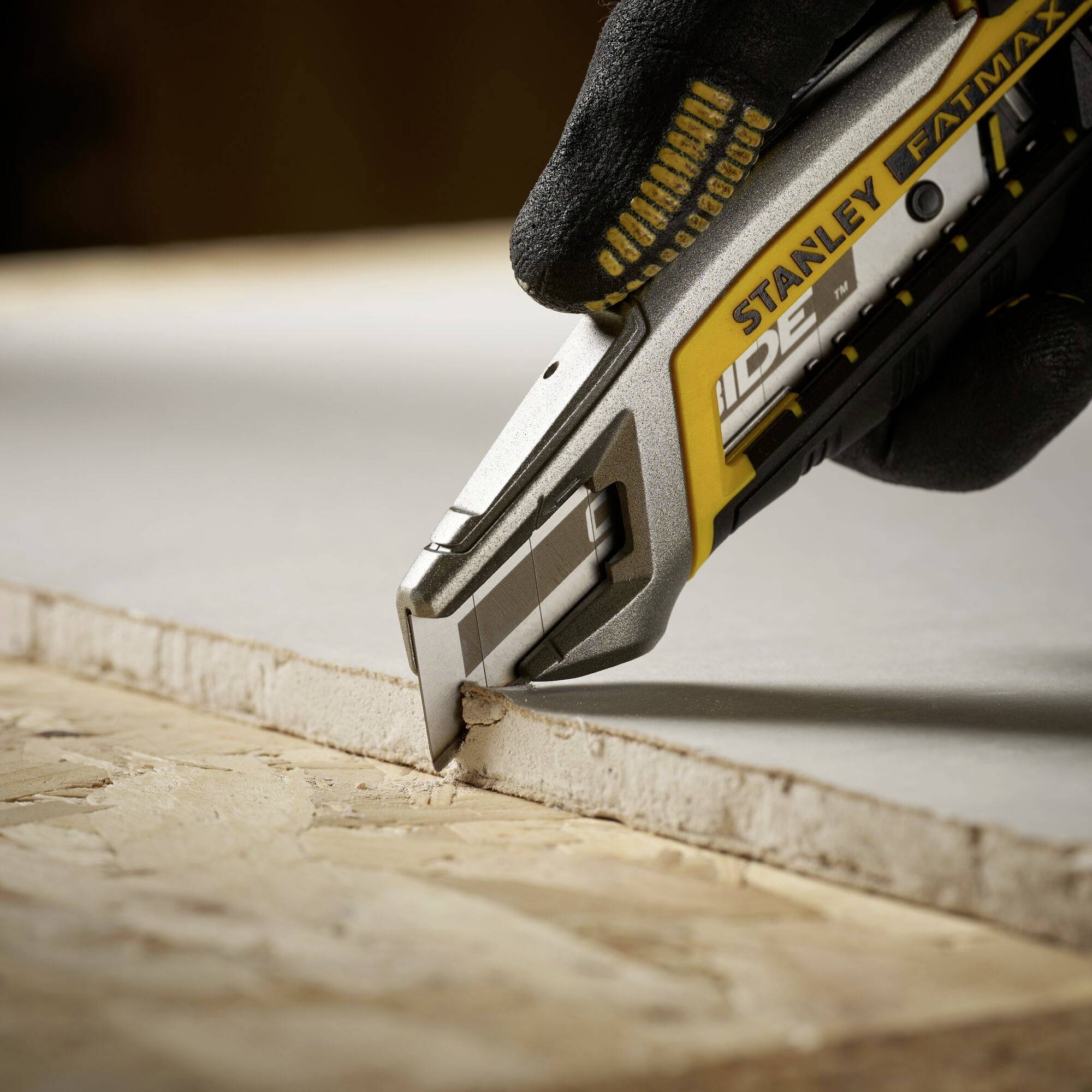 Une main coupe une plaque de plâtre avec un couteau Stanley FatMax. Le couteau a une couleur jaune et grise et une lame robuste.