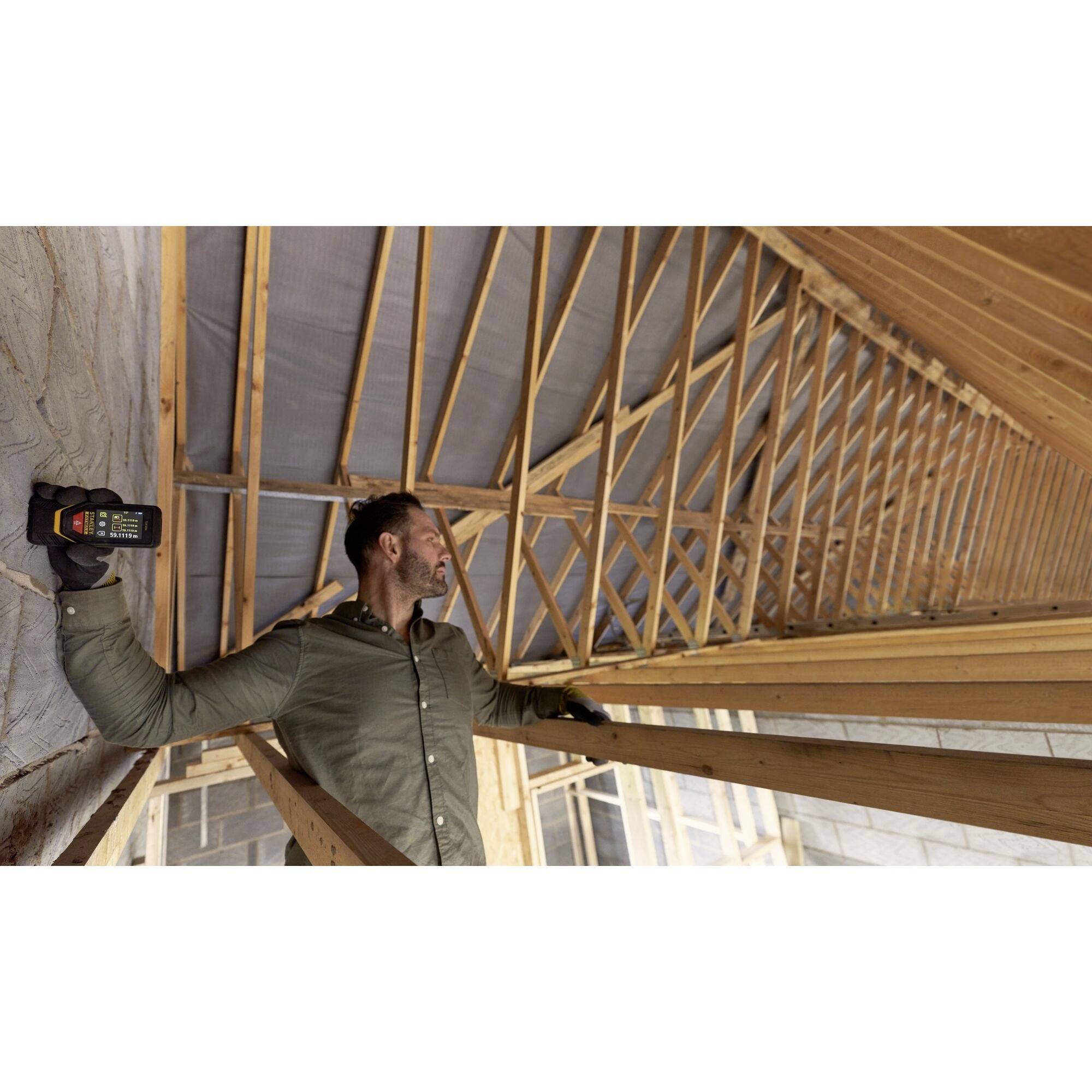Un homme sur un chantier vérifie l'intérieur d'une charpente en bois à l'aide d'un appareil de mesure. Il porte des vêtements chauds et des gants.