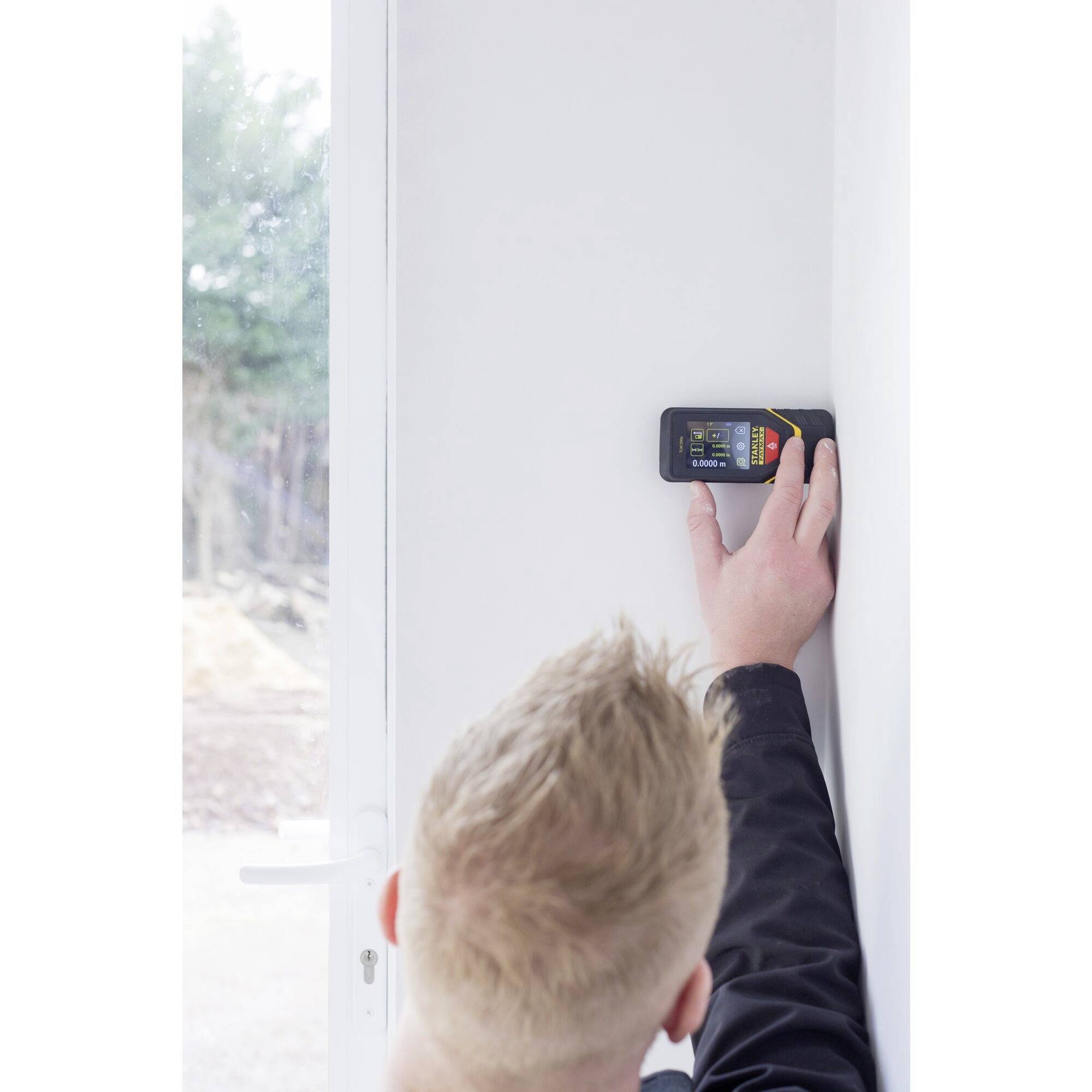 Une personne mesure la distance sur un mur blanc à l'aide d'un appareil numérique, entourée d'un encadrement de fenêtre avec vue sur l'extérieur.