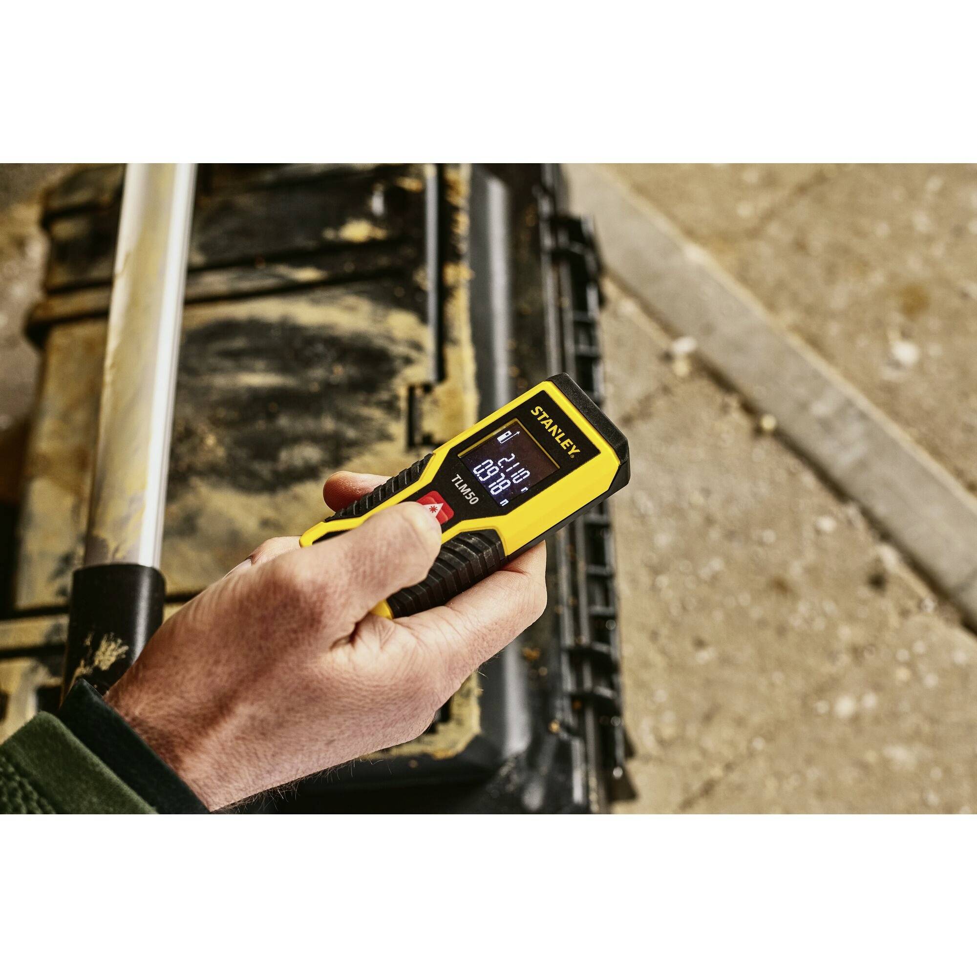 Une personne utilise un télémètre laser numérique jaune pour mesurer une distance de 4,234 mètres. Une boîte à outils est visible en arrière-plan.