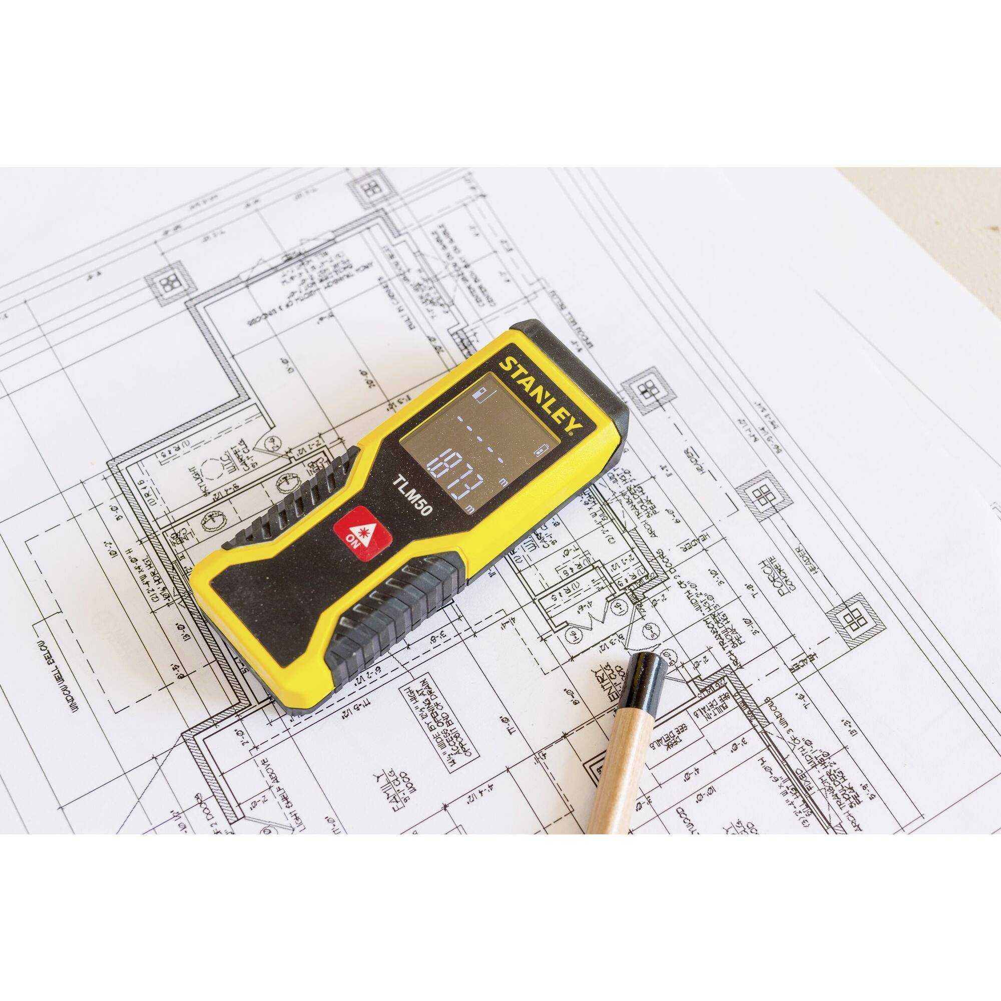 Un appareil de mesure jaune est posé sur un plan d'architecture, à côté duquel se trouve un crayon. L'affichage indique '1.79'. L'accent est mis sur la précision de la mesure.