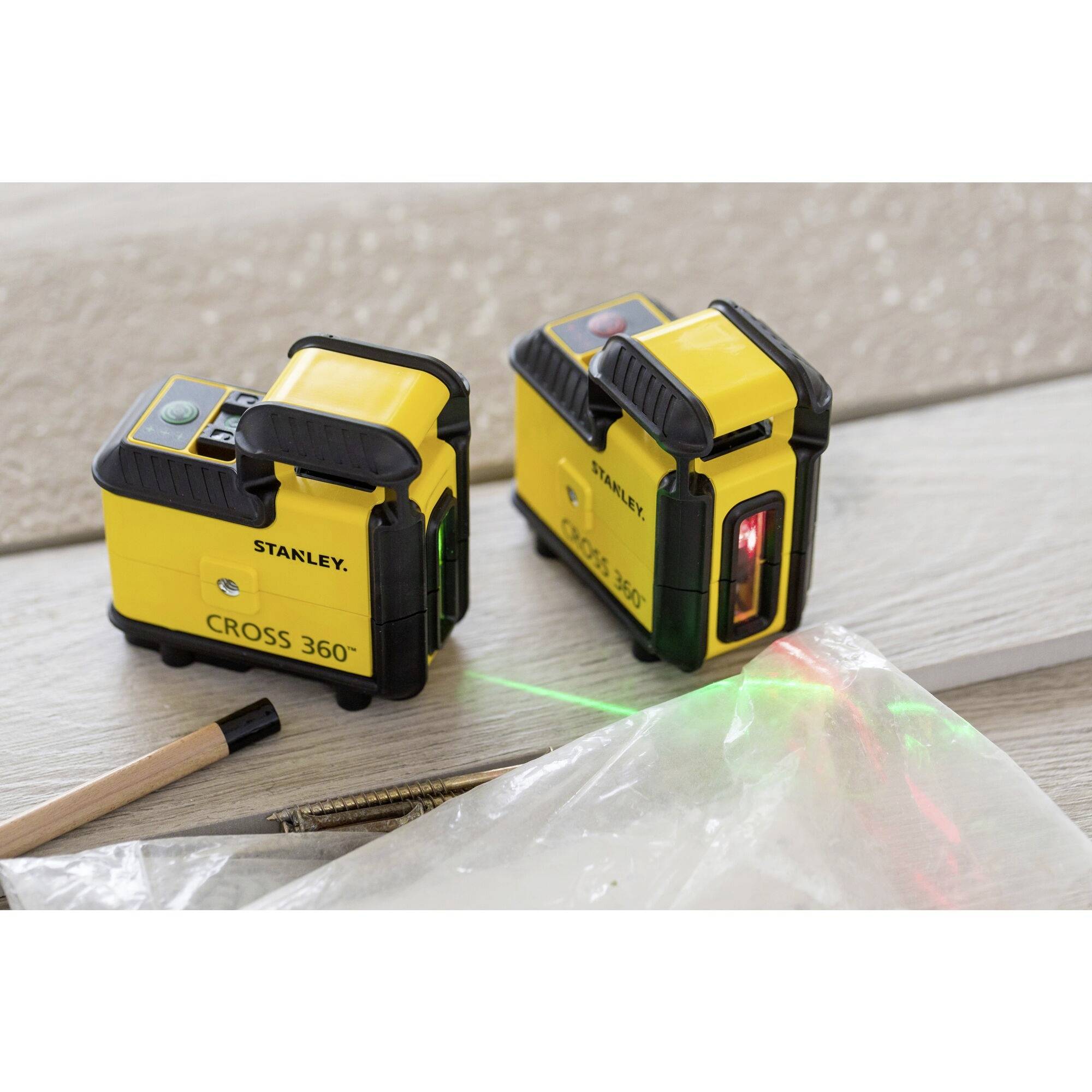 Deux niveaux laser jaunes sur un sol en bois ; l'un projette des lignes rouges et vertes. Au premier plan, un crayon et des vis.