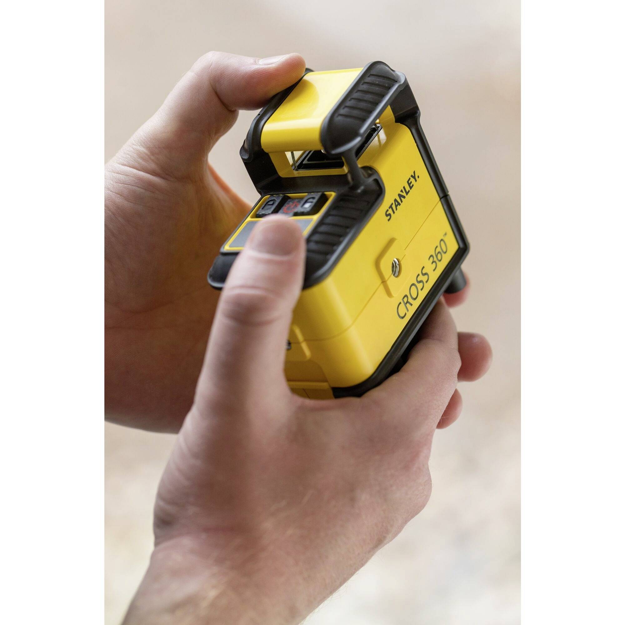 Une personne tient un appareil jaune carré portant l'inscription 'CROSS 360' et le nom de la marque. L'appareil semble être un outil.