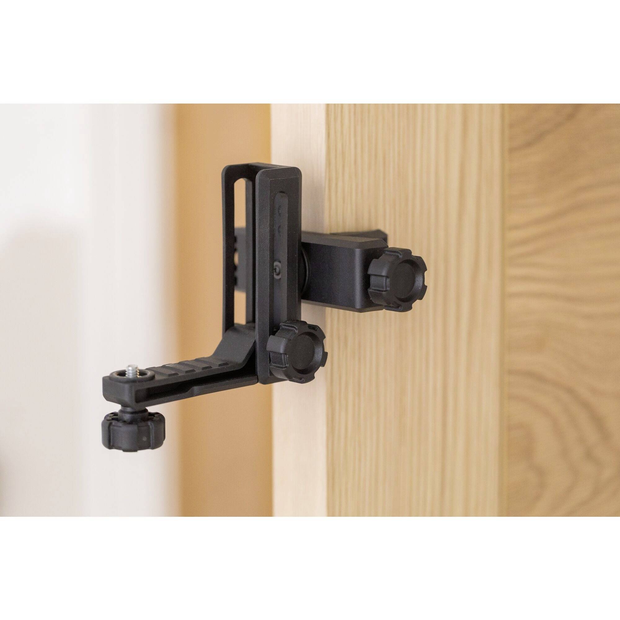Un arrêt-porte noir est fixé à une porte en bois. Le support comporte plusieurs boutons rotatifs réglables pour le maintien sur la porte.