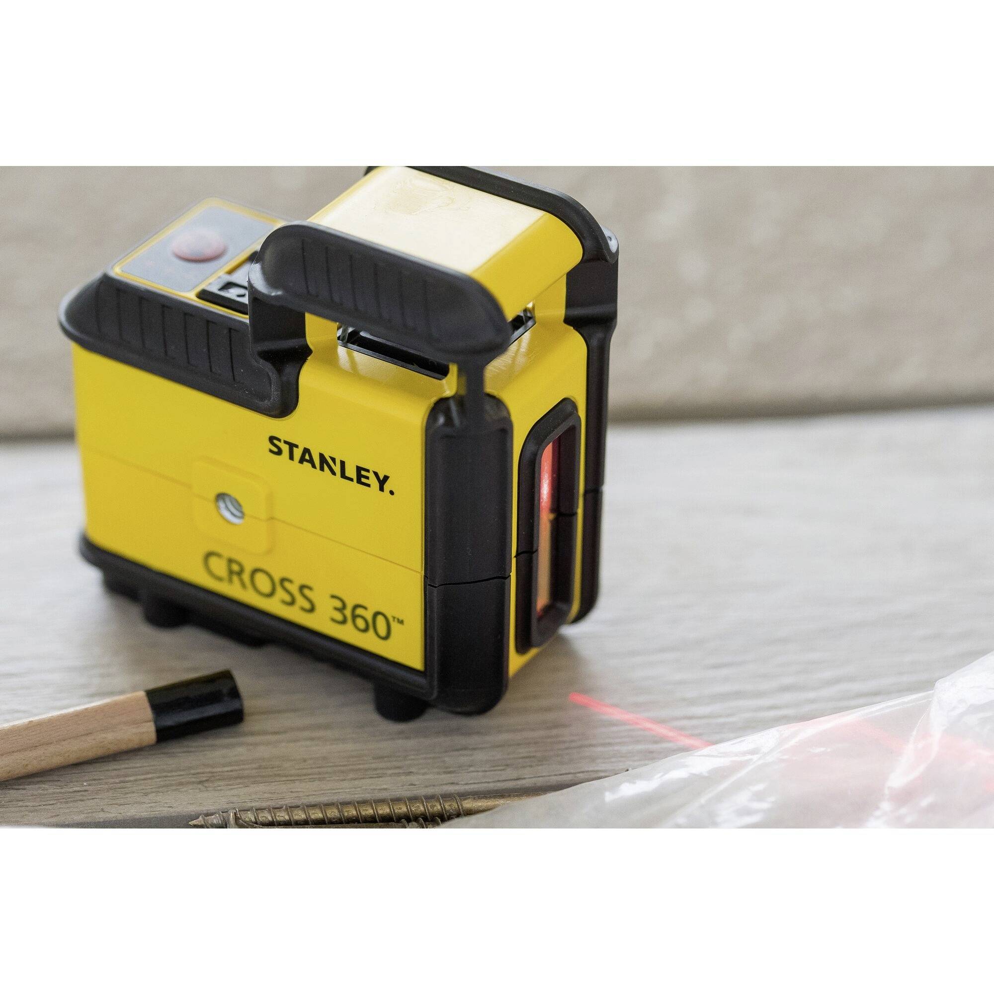 Un appareil laser à lignes croisées jaune portant l'inscription 'Stanley Cross 360°', positionné sur un sol en bois. Un stylo et des vis sont disposés à côté.