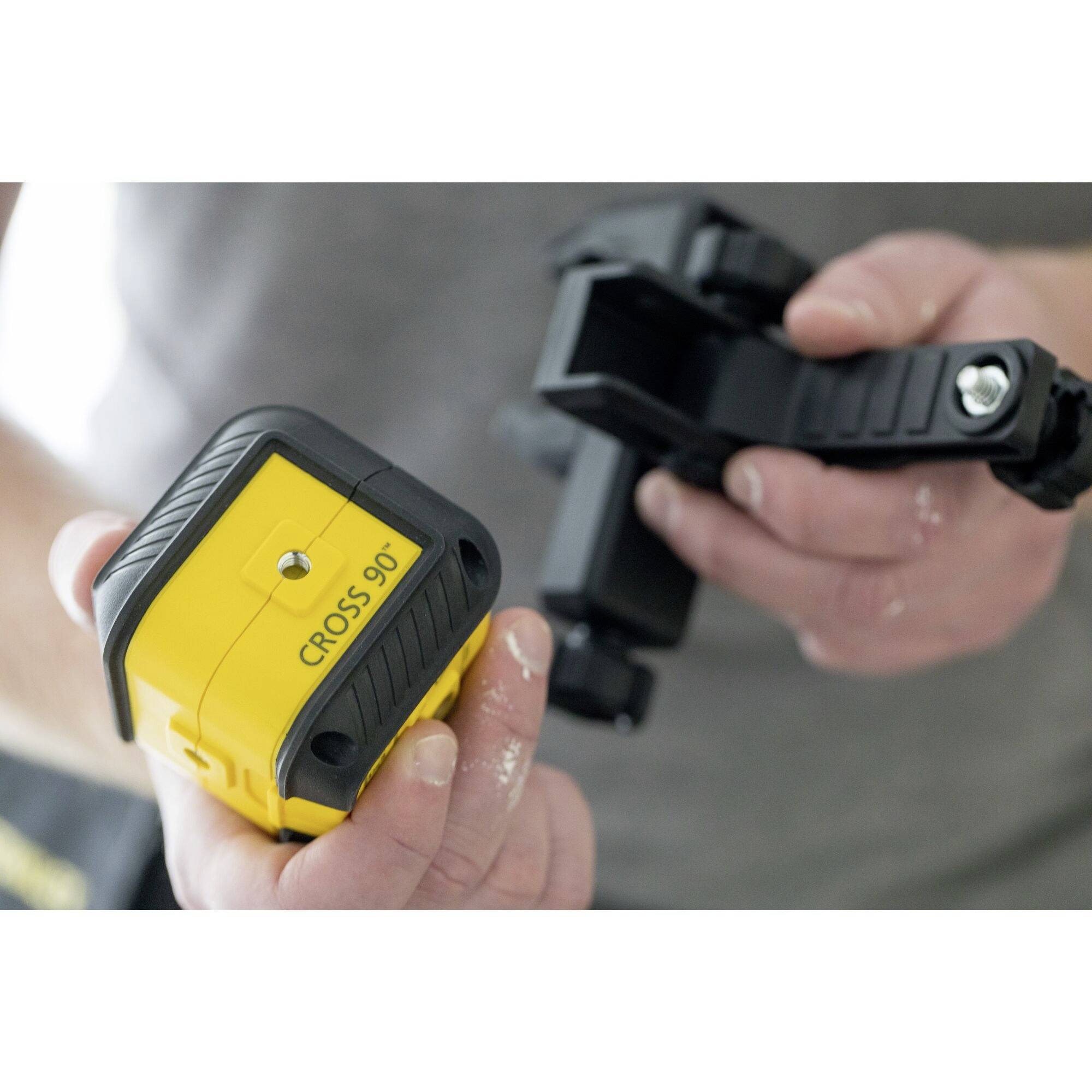 Une personne tient un outil de mesure jaune portant l'inscription 'CROSS 90°', fixé à un support noir. La mise au point est sur l'appareil.