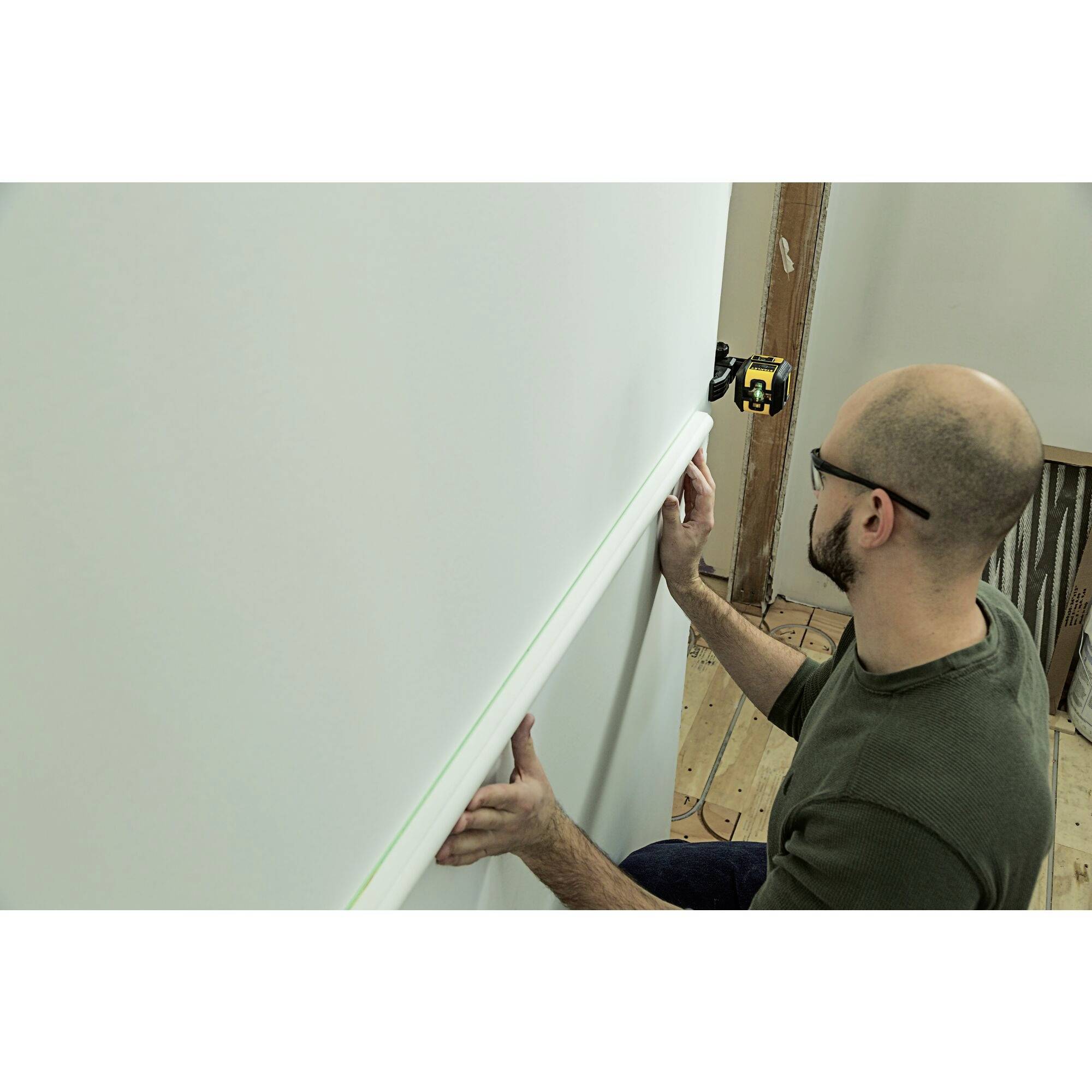 Un homme installe une corniche blanche sur un mur en utilisant un dispositif de mesure laser pour un alignement précis.