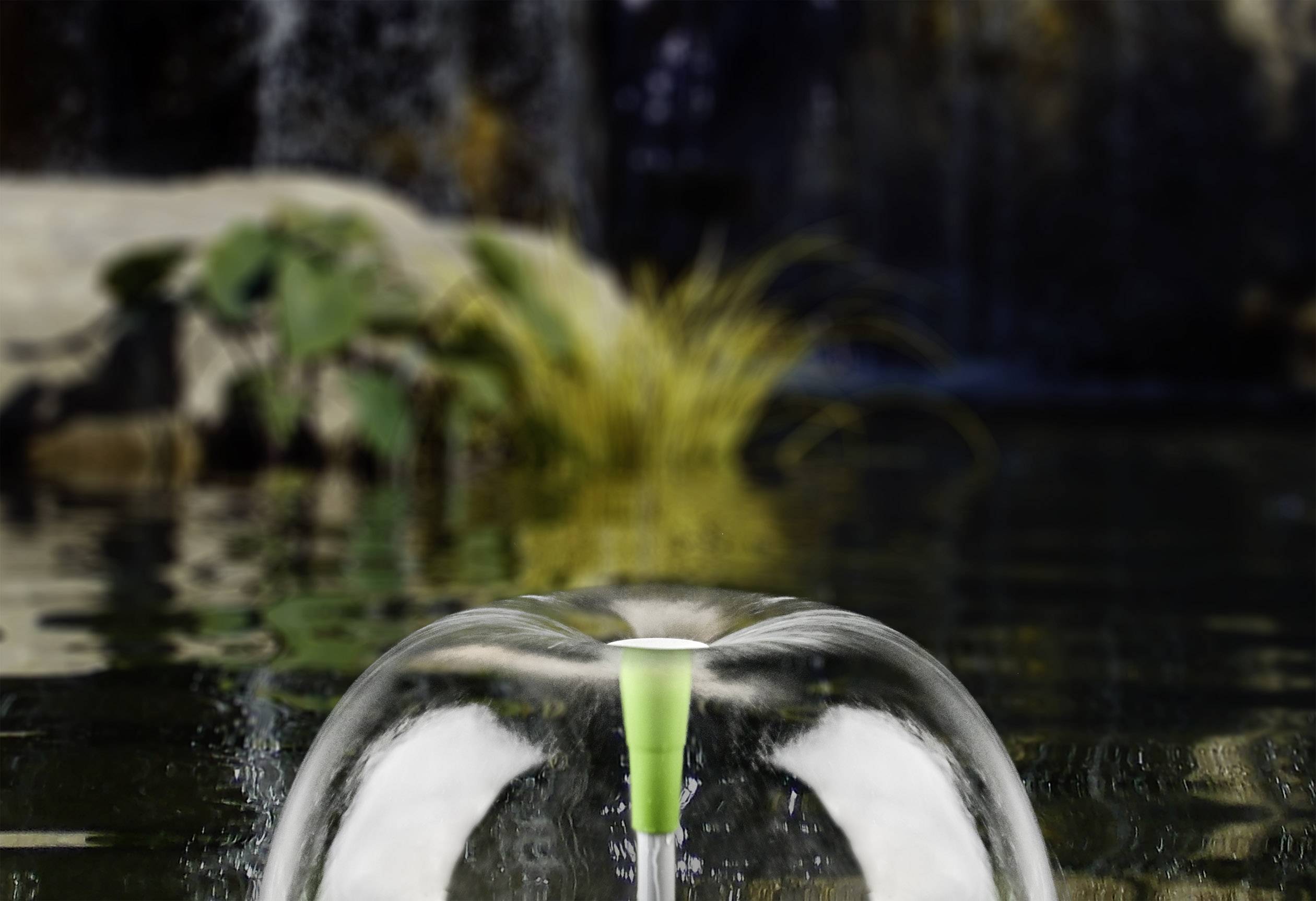 Une fontaine dans l'eau forme un dôme hémisphérique clair. En arrière-plan, on aperçoit des plantes et une cascade.